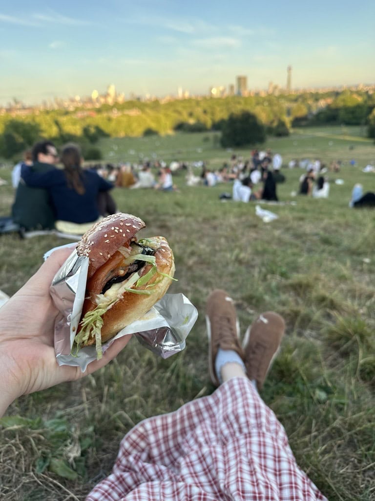 Someone holding up a burger with a view of Primrose Hill London in the background.