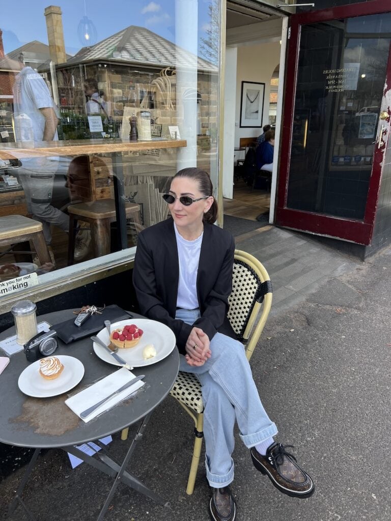 Emma Jackman, ROMY founder, seated at an outdoor café table with pastries, wearing sunglasses, a black blazer, white tee, and wide-leg jeans.
