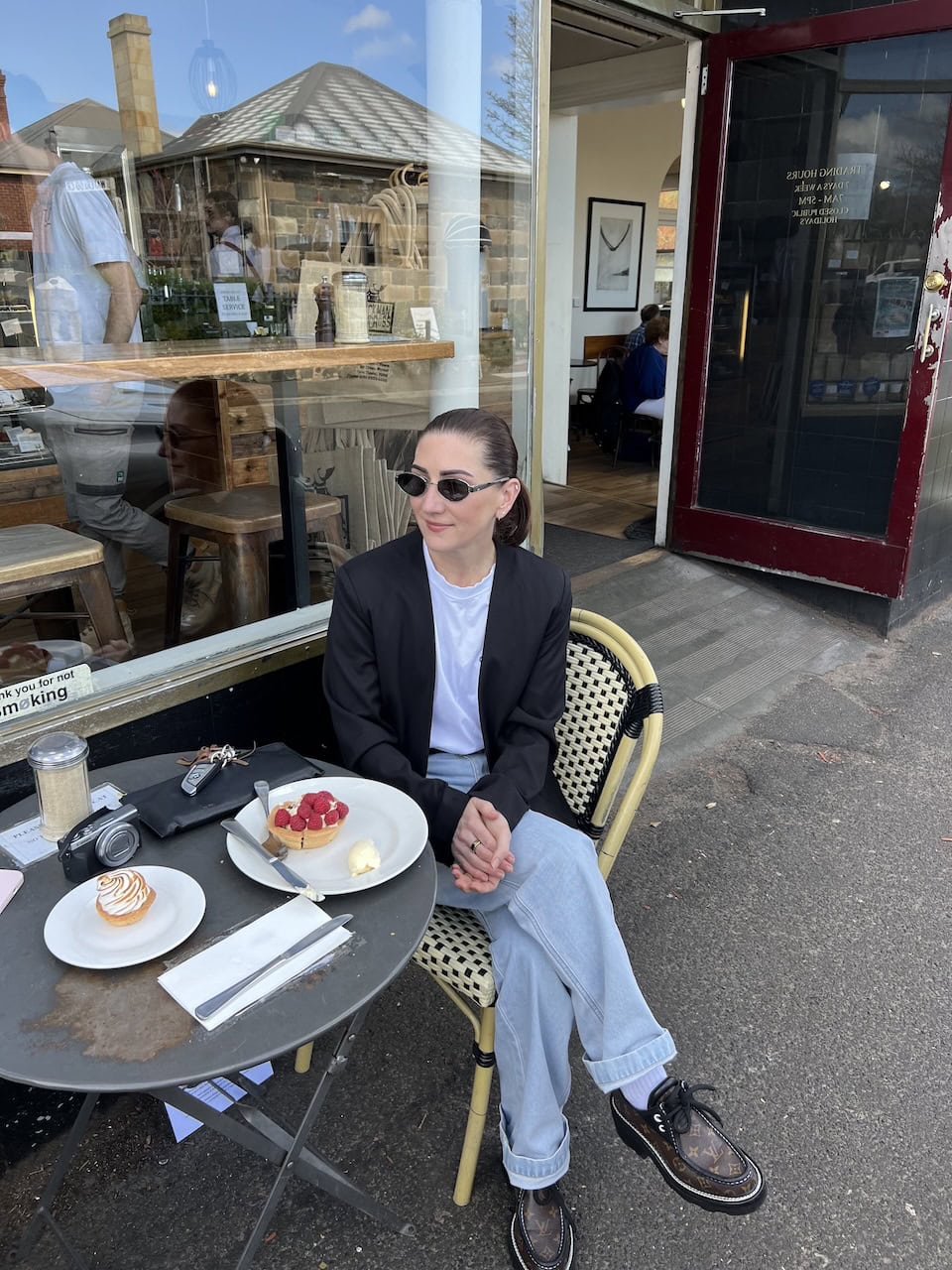Emma Jackman, ROMY founder, seated at an outdoor café table with pastries, wearing sunglasses, a black blazer, white tee, and wide-leg jeans.