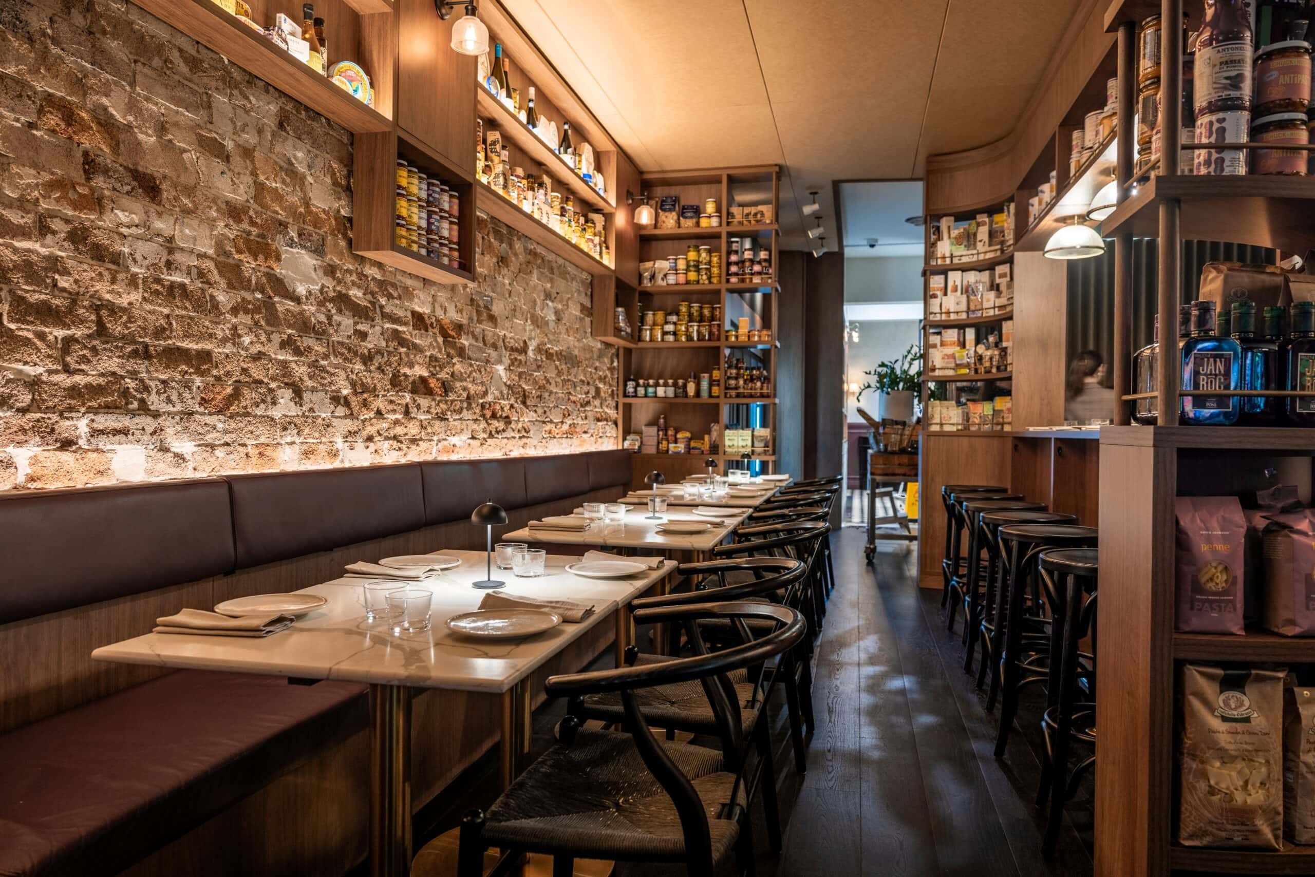 Exposed sandstone brick wall with warm-lit deli shelving banquette seating and marble tables at Duke's Providore Paddington Sydney