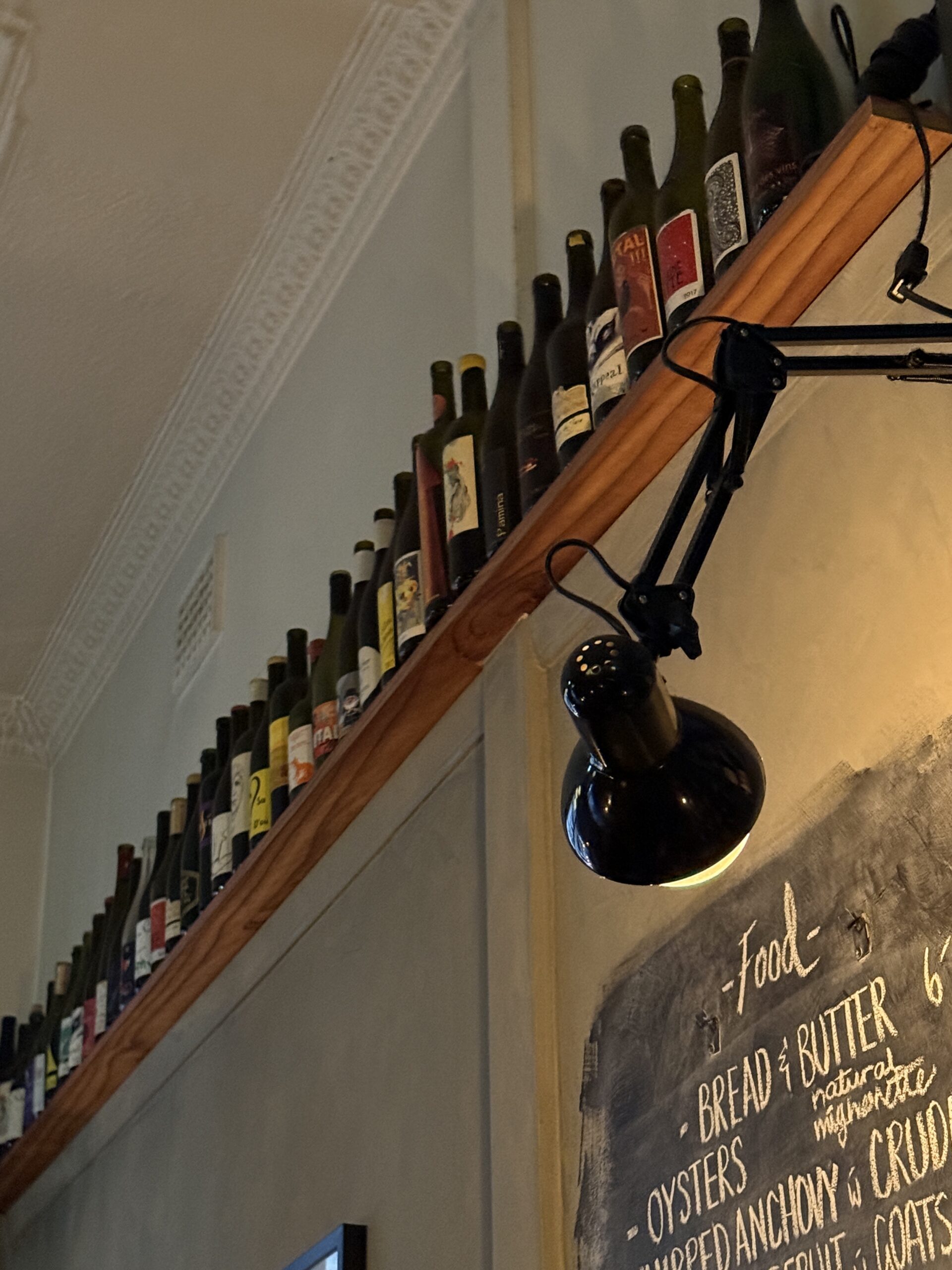 A timber shelf lined with natural wine bottles at Lucinda Wine Bar, with a black articulated lamp and handwritten chalkboard menu below.