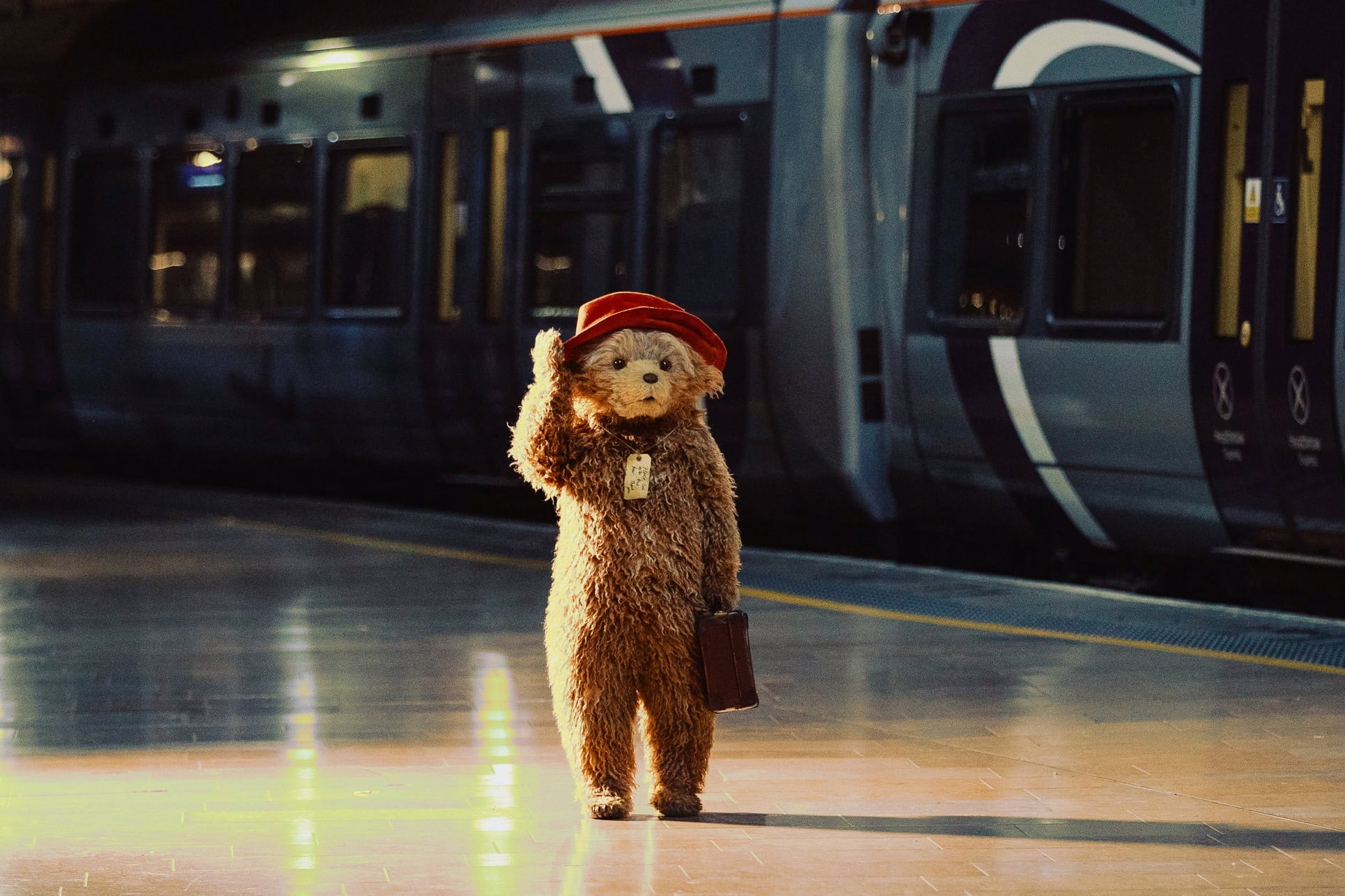 Paddington Bear waving on a train platform with a suitcase and red hat for Paddington the Musical
