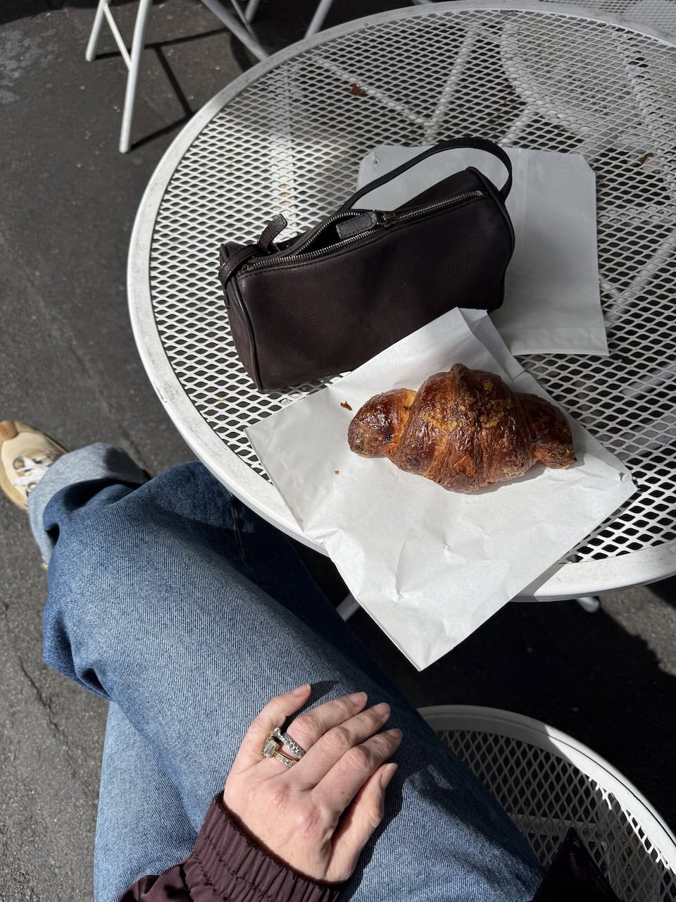 A golden croissant on baking paper beside a dark leather handbag on an outdoor mesh café table at Queens Bakery.
