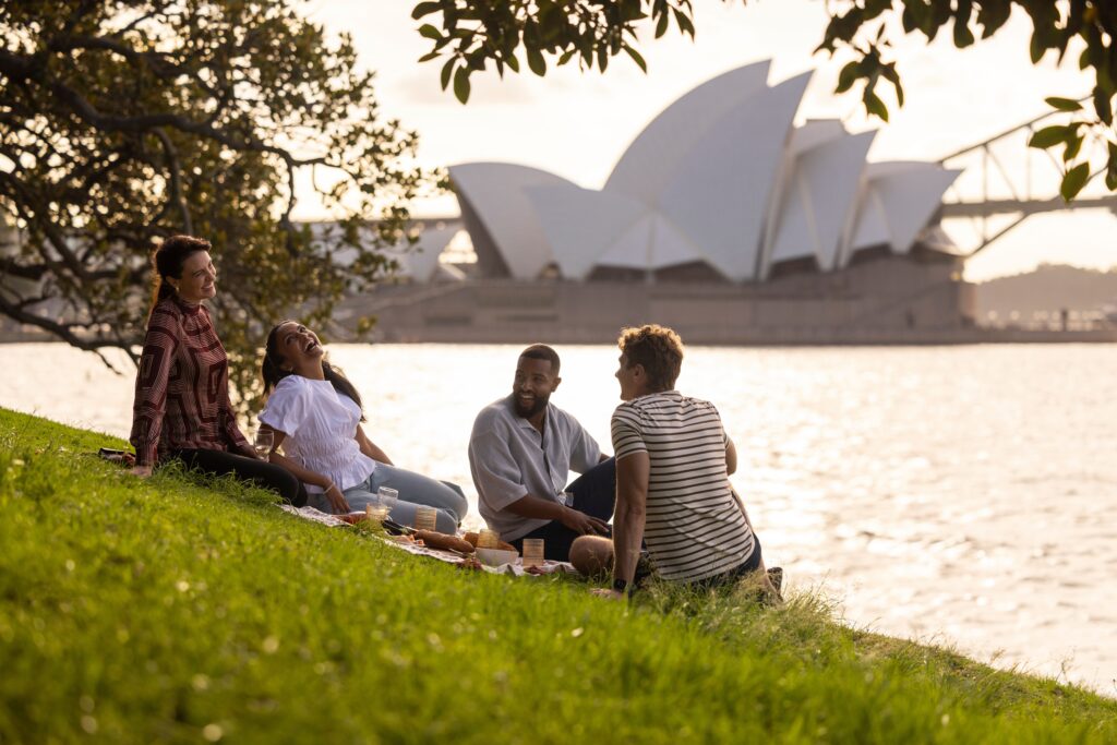 A group of friends enjoying a picnic at the royal botanic gardens in Sydney with the Sydney Opera House in the background.