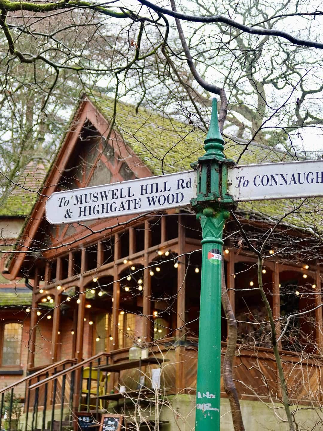 A green and white signpost out the front of the fairytale-like and festoon light-strewn Queen's Wood Cafe in Highgate.