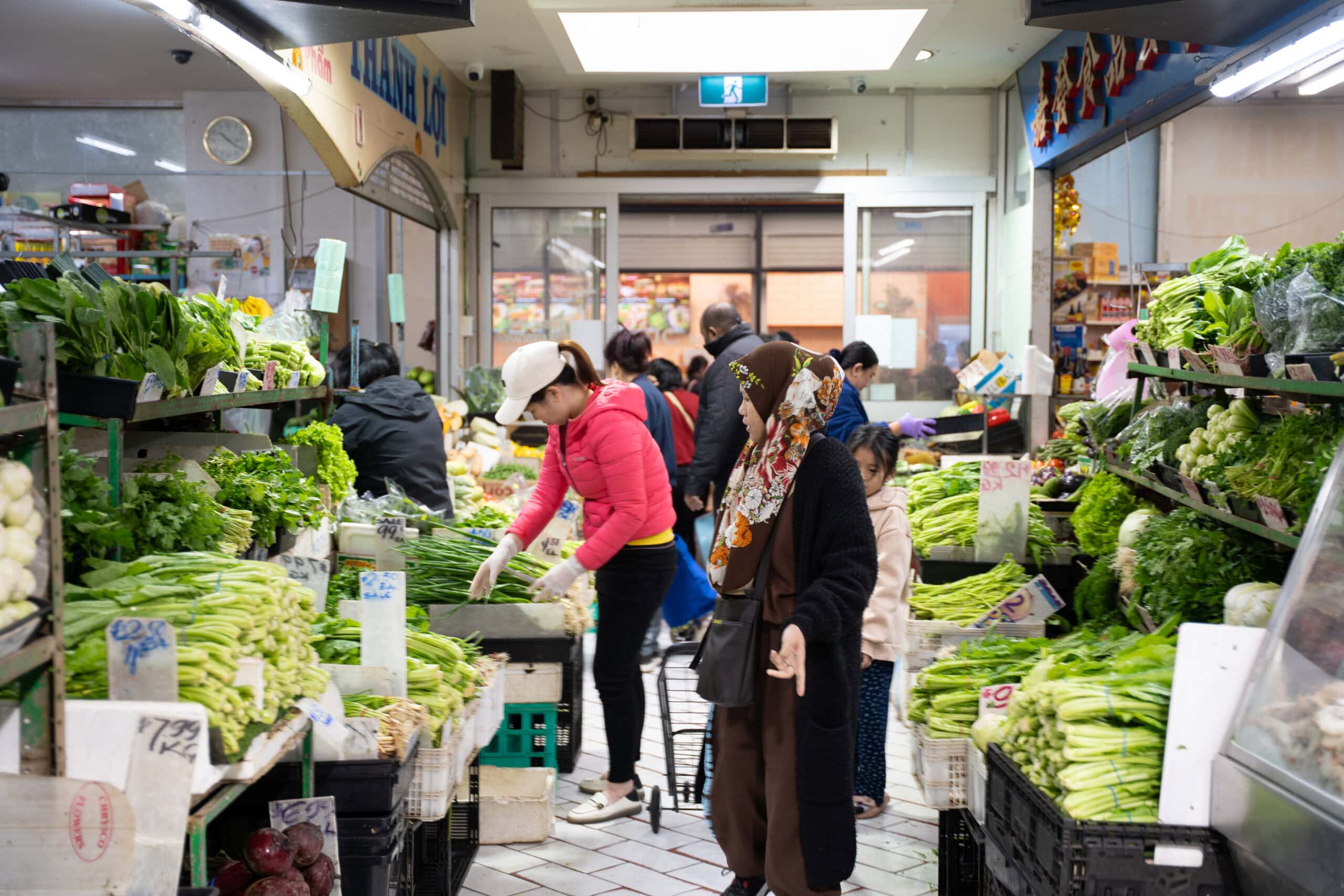 Indoor market with shoppers browsing leafy greens and Asian vegetables.