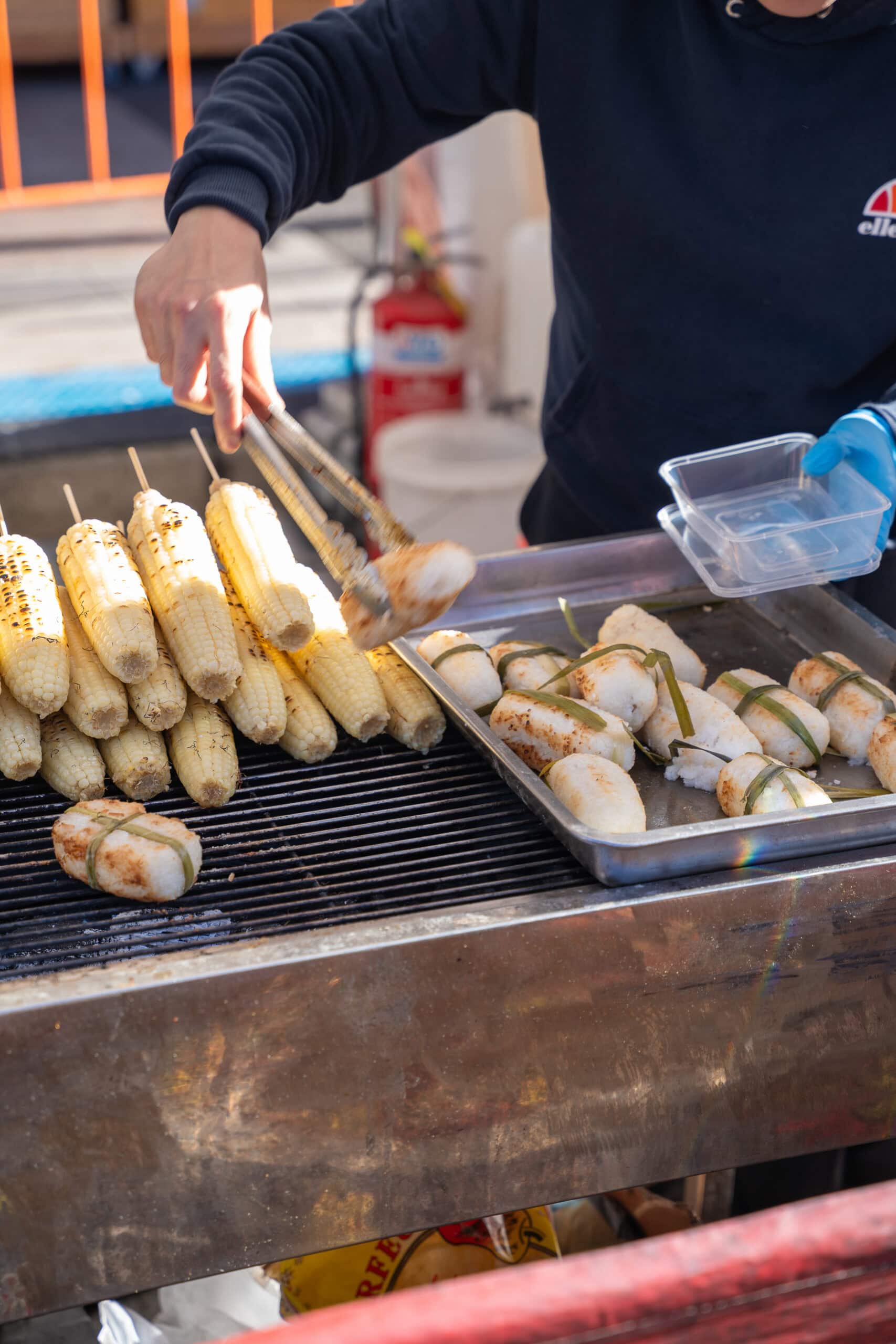 Vendor grilling corn and banana leaf-wrapped sticky rice at a festival food stall.