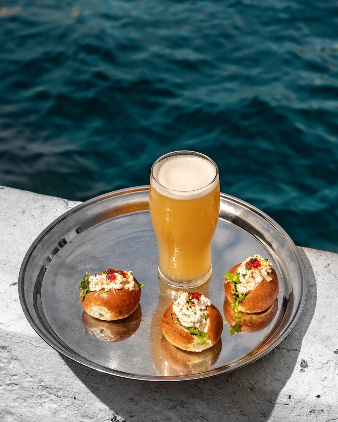 A silver tray with lobster rolls and a beer on it, placed next to the water at Cruise Bar in Circular Quay.