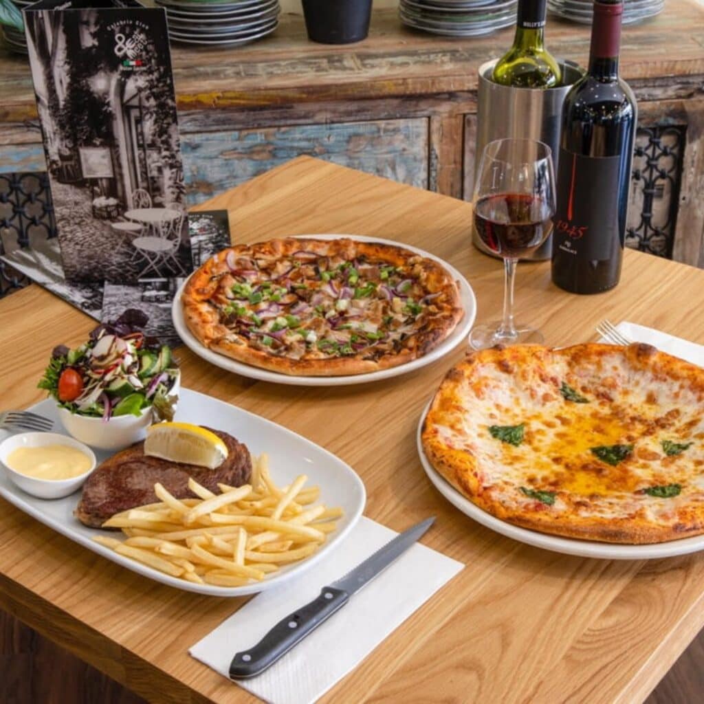 A square wooden table with two pizzas and a steak and chips on top.