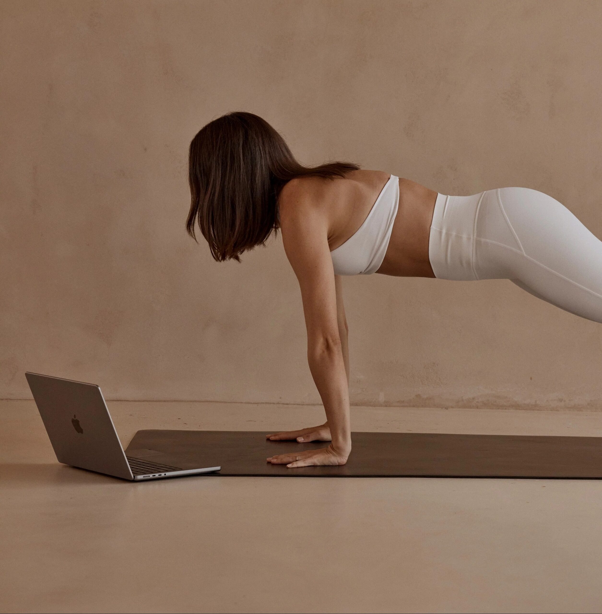 Woman in plank pose on yoga mat with laptop following online pilates class