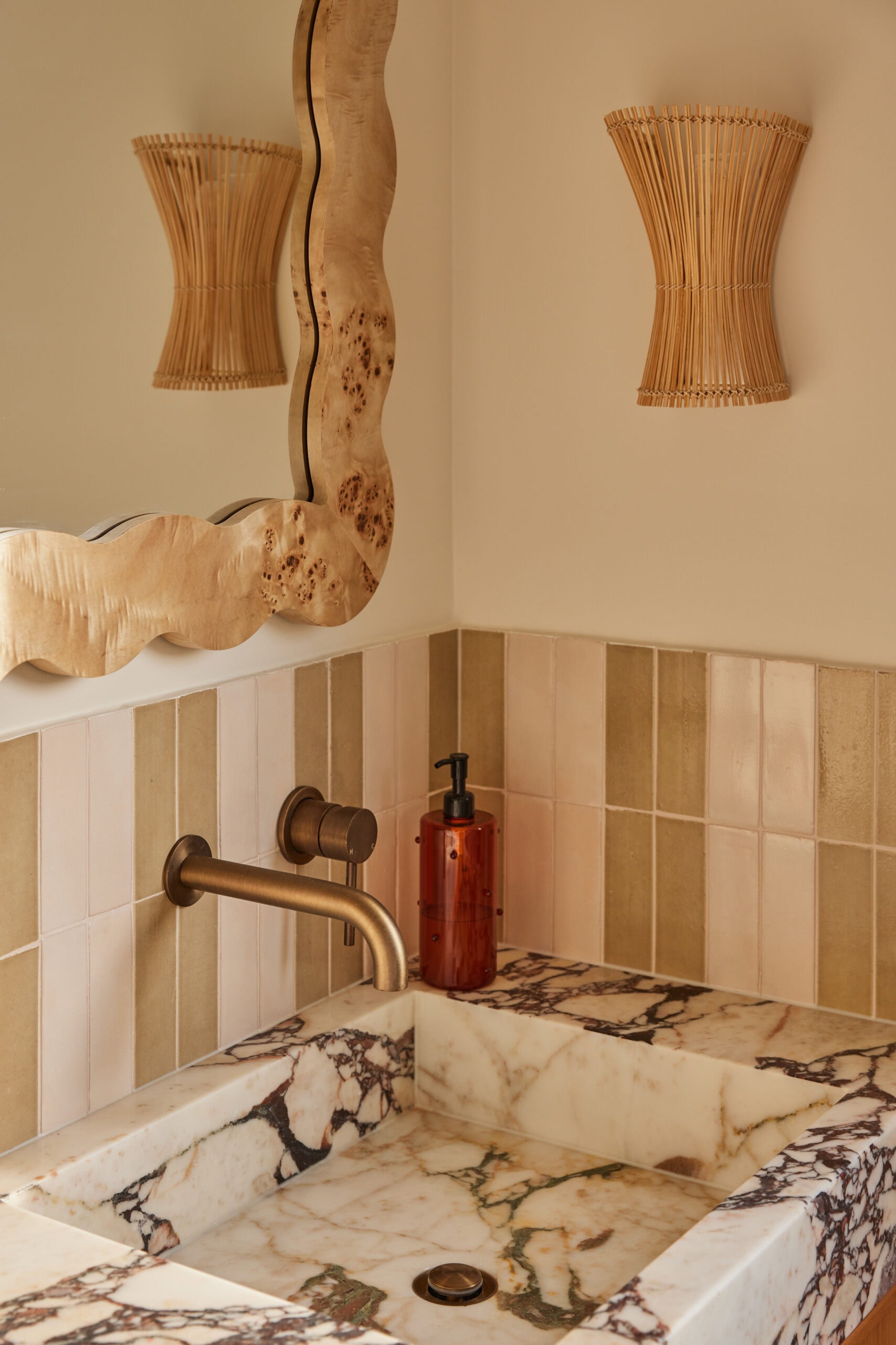 Wavy burl wood mirror reflecting a woven rattan wall sconce above a veined marble vanity, with a checkerboard trinket box and green and cream striped tiles.