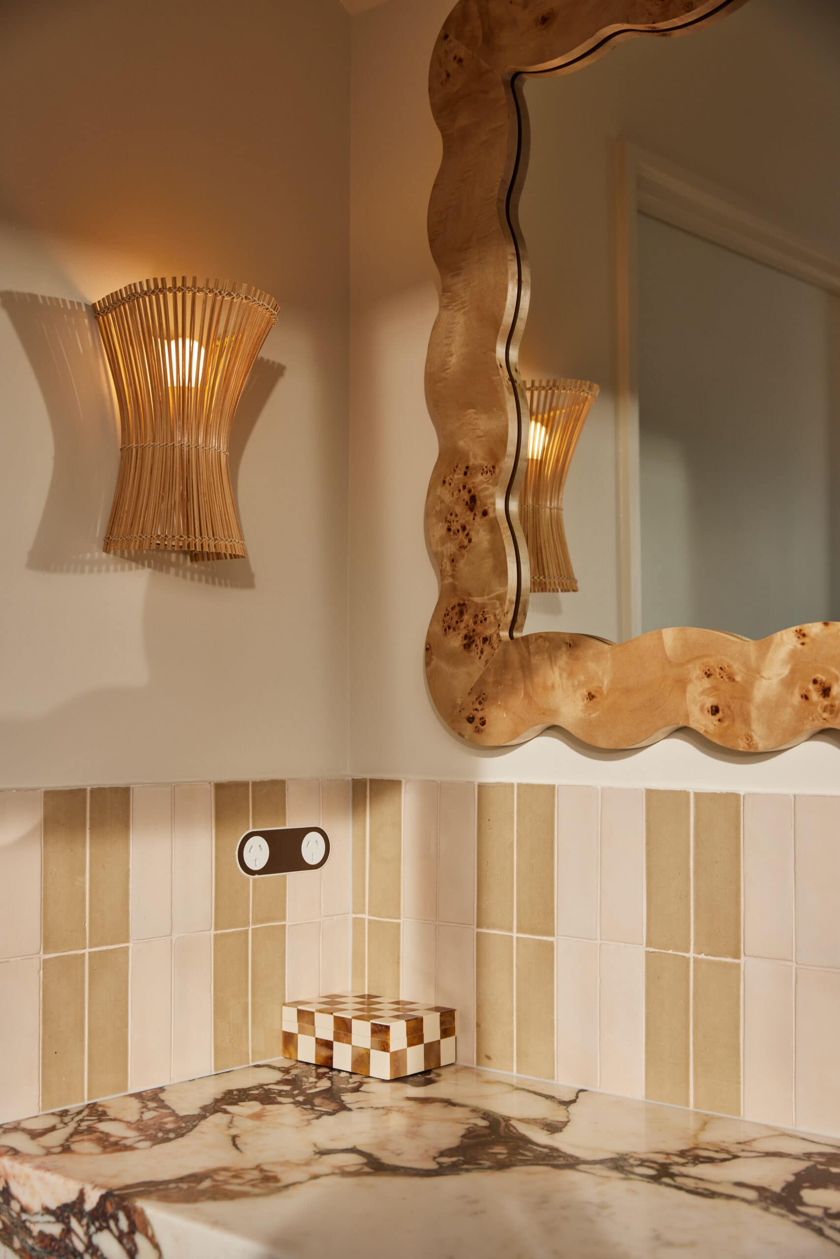 Carved marble corner sink with an antique brass wall-mounted tap, an amber glass soap pump, woven rattan wall sconces, and a wavy burl wood mirror against green and pink striped tiles.