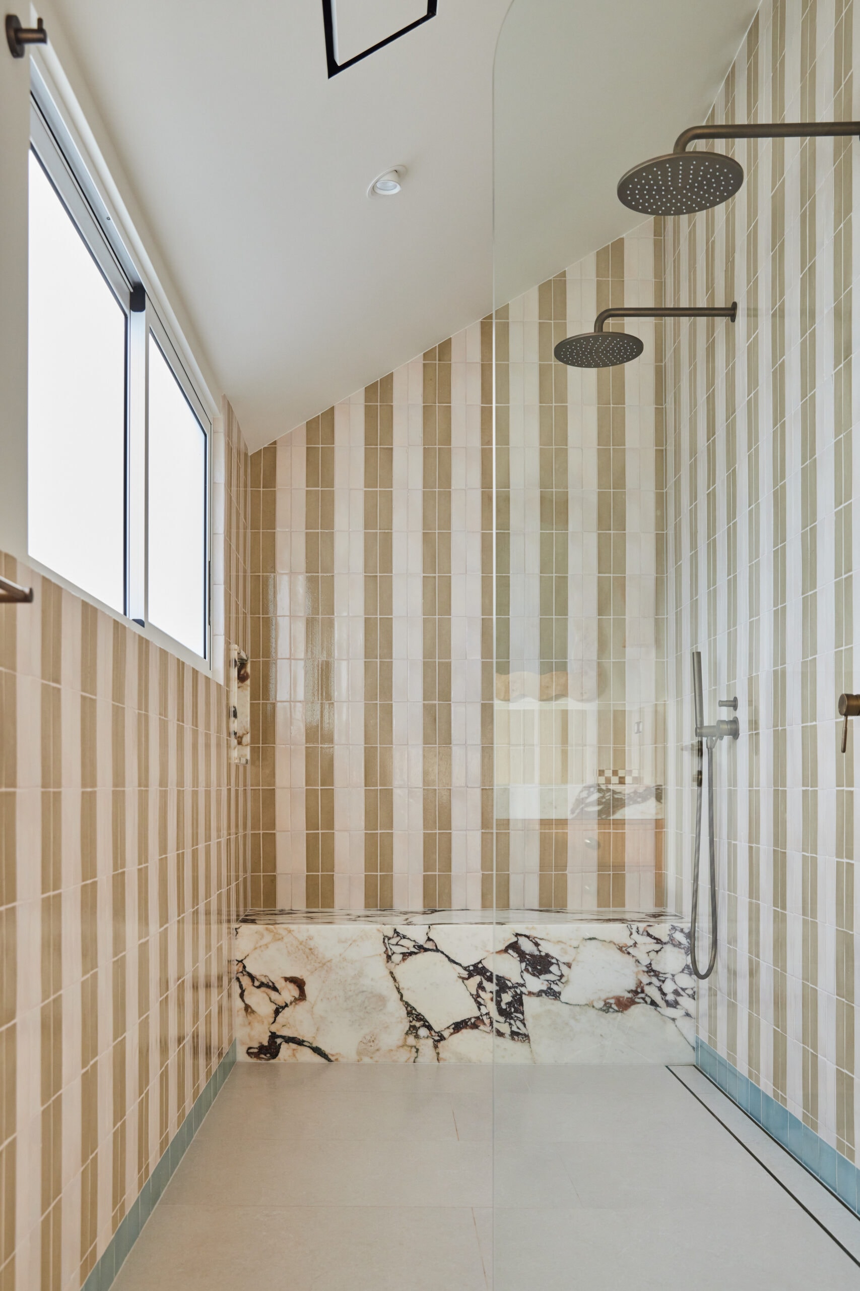 Walk-in shower with green and cream striped tiles, a veined marble seat, dual antique brass rain showerheads, a handheld shower attachment, and a frameless glass screen.