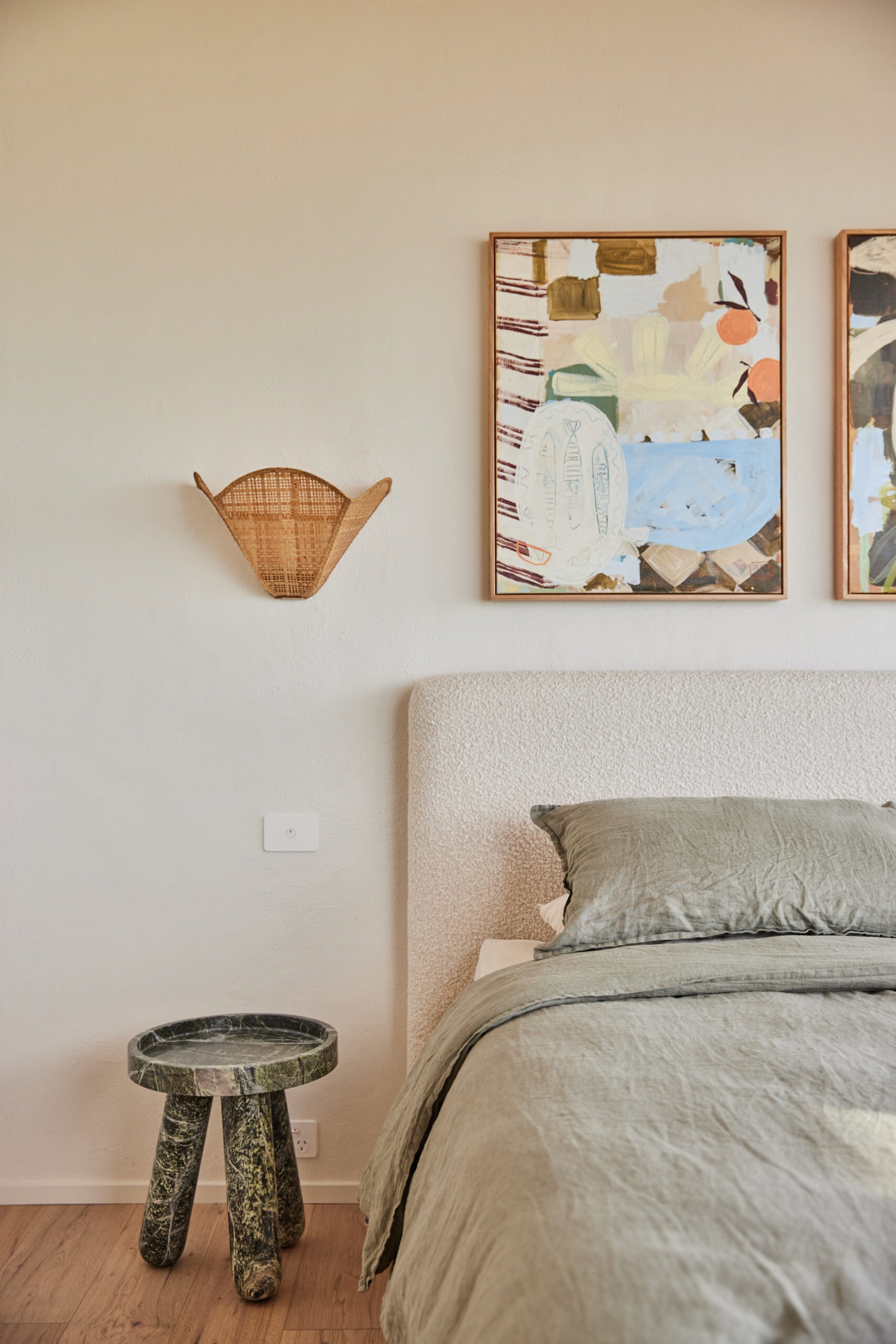 Bedroom with sage green bedding under colourful artwork, rattan wall sconce and marble side table.