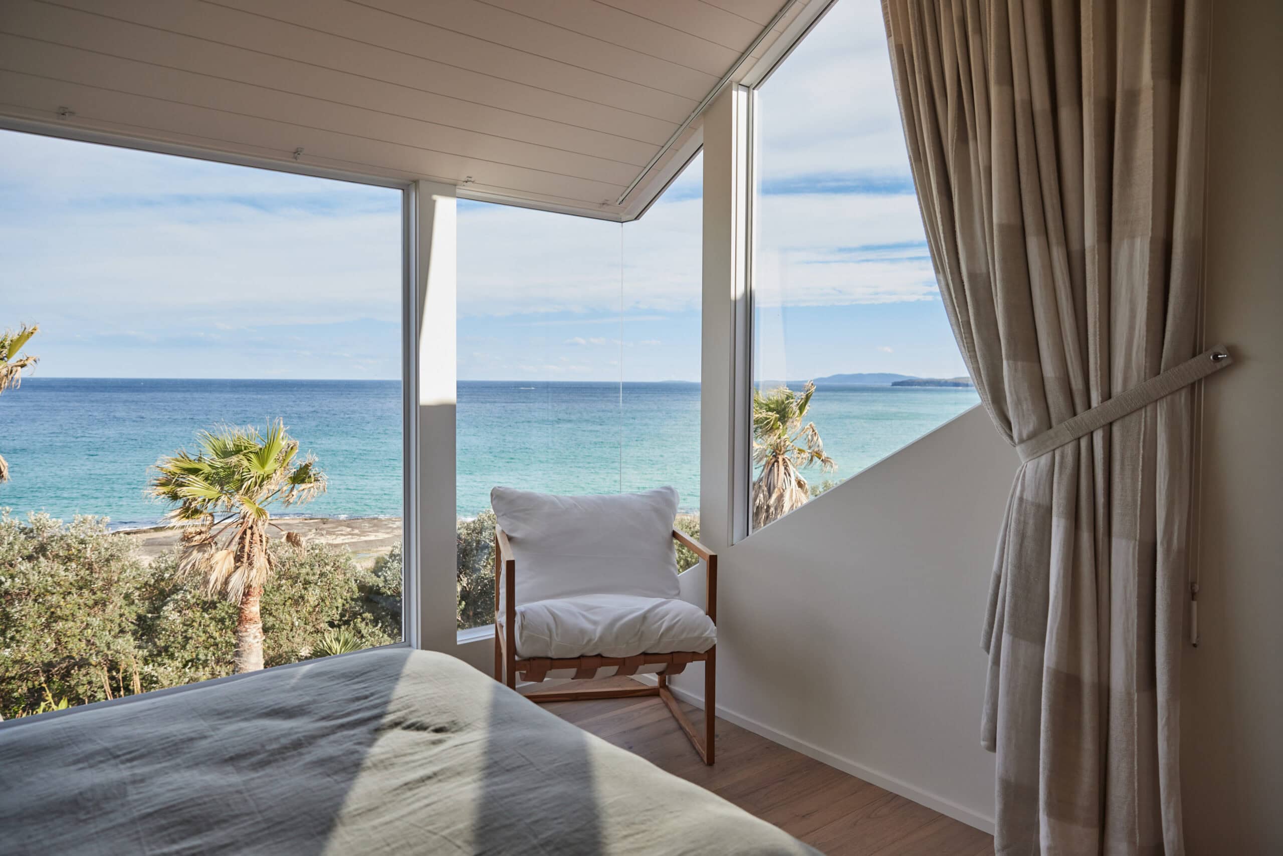 Bedroom corner with an angled ceiling and floor-to-ceiling windows framing an ocean view, a linen armchair by the window, and pleated curtains tied back.