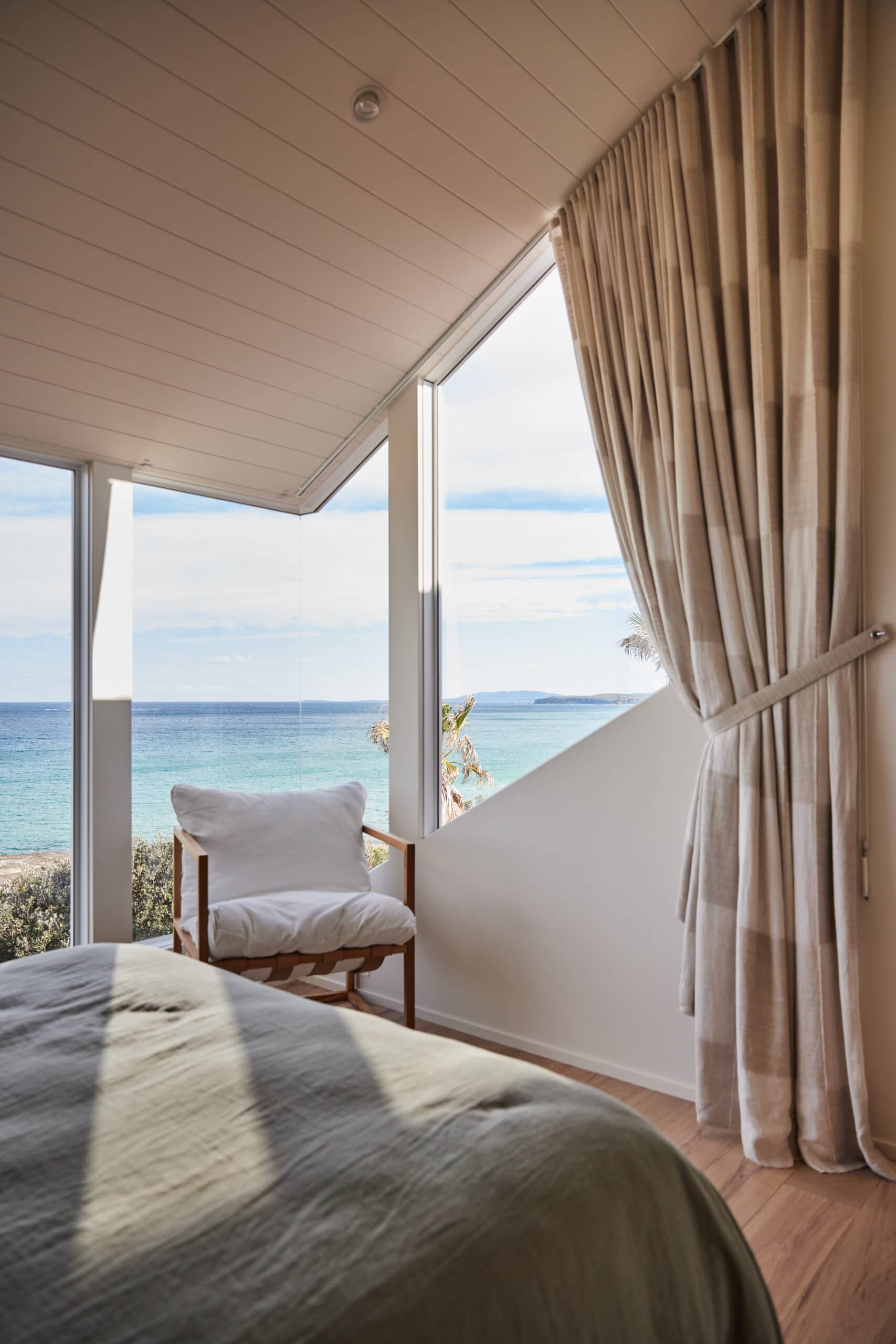Bedroom nook with a wraparound window looking out over turquoise ocean water, palm trees, and a beach, with a linen armchair positioned to take in the view.