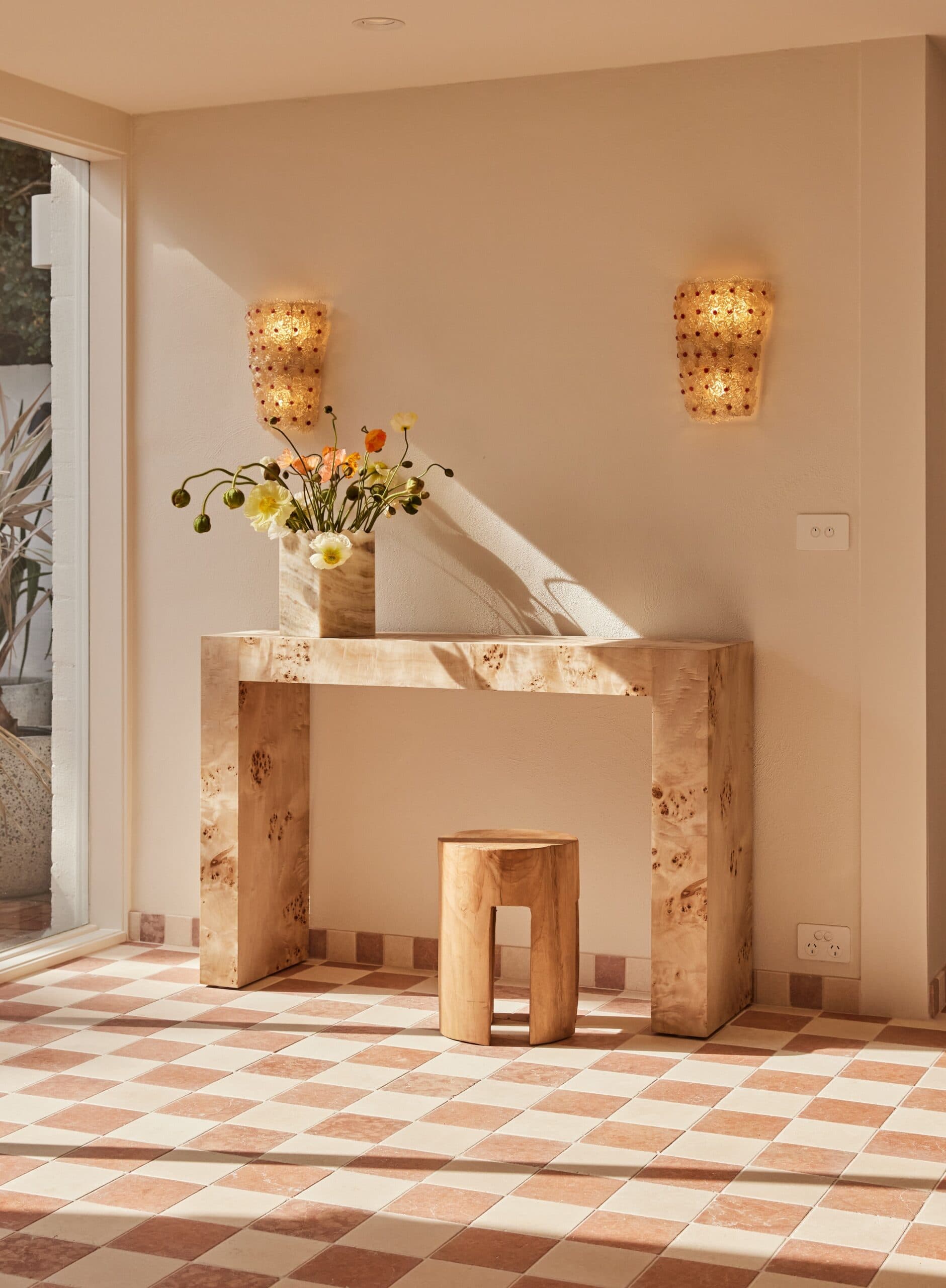 Sunlit hallway with a burl wood console table, travertine vase of poppies, scalloped glass wall sconces, and a round timber stool on terracotta checkerboard tile floor.