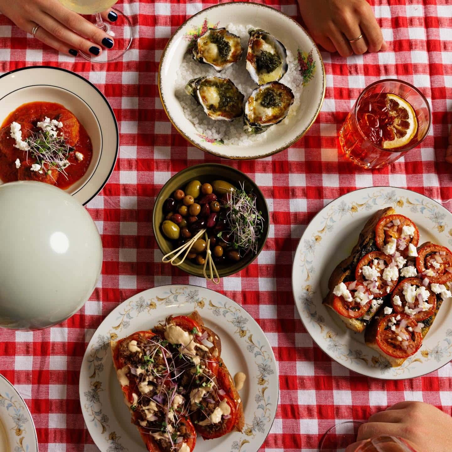 Overhead spread of oysters, tomato toast, olives and cocktails on red gingham tablecloths at Holly’s in Ringwood.