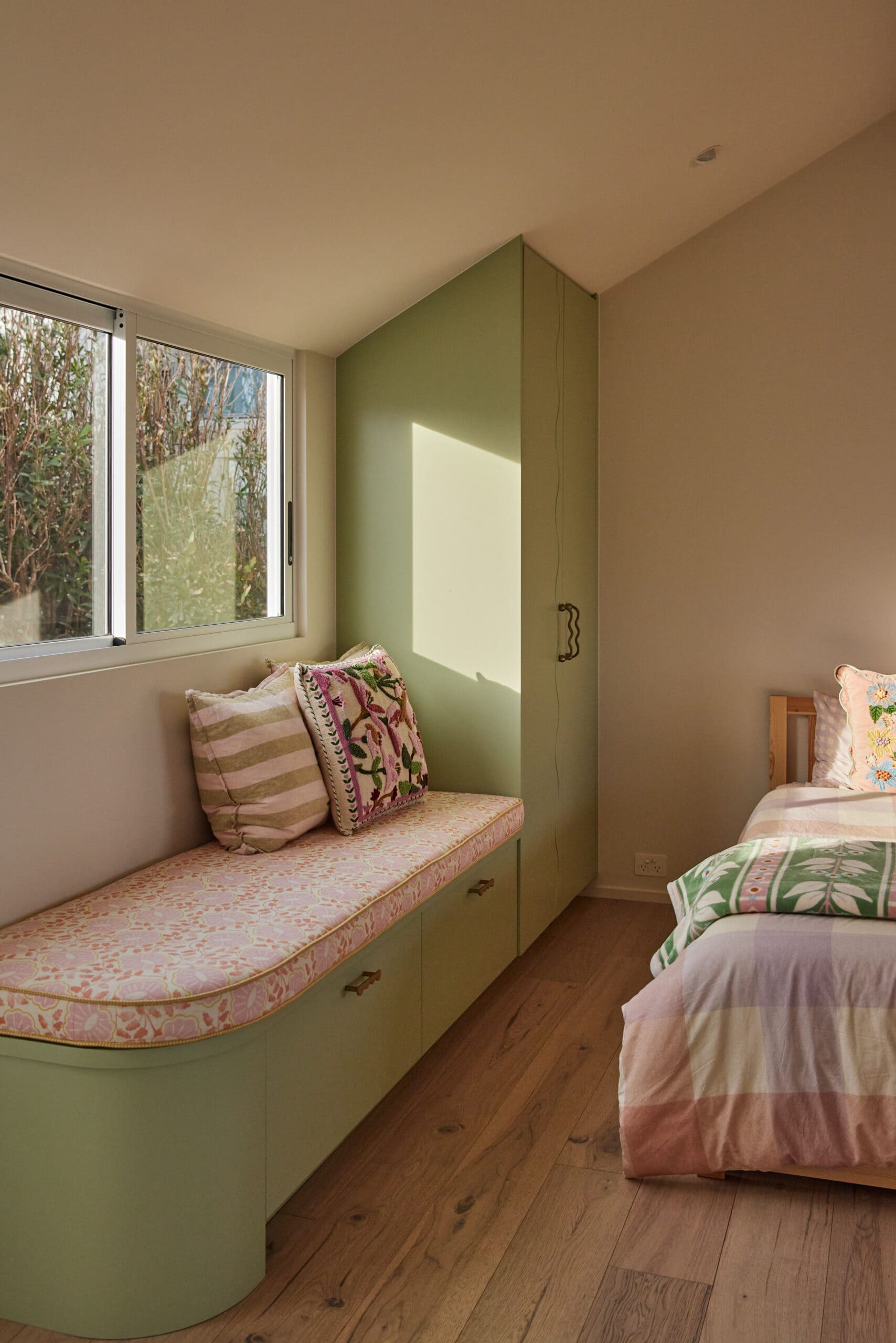 Kids' bedroom with a sage green built-in window seat and wardrobe, floral and striped throw pillows, a pink patterned cushion, and a twin bed dressed in pink and green patterned linens.