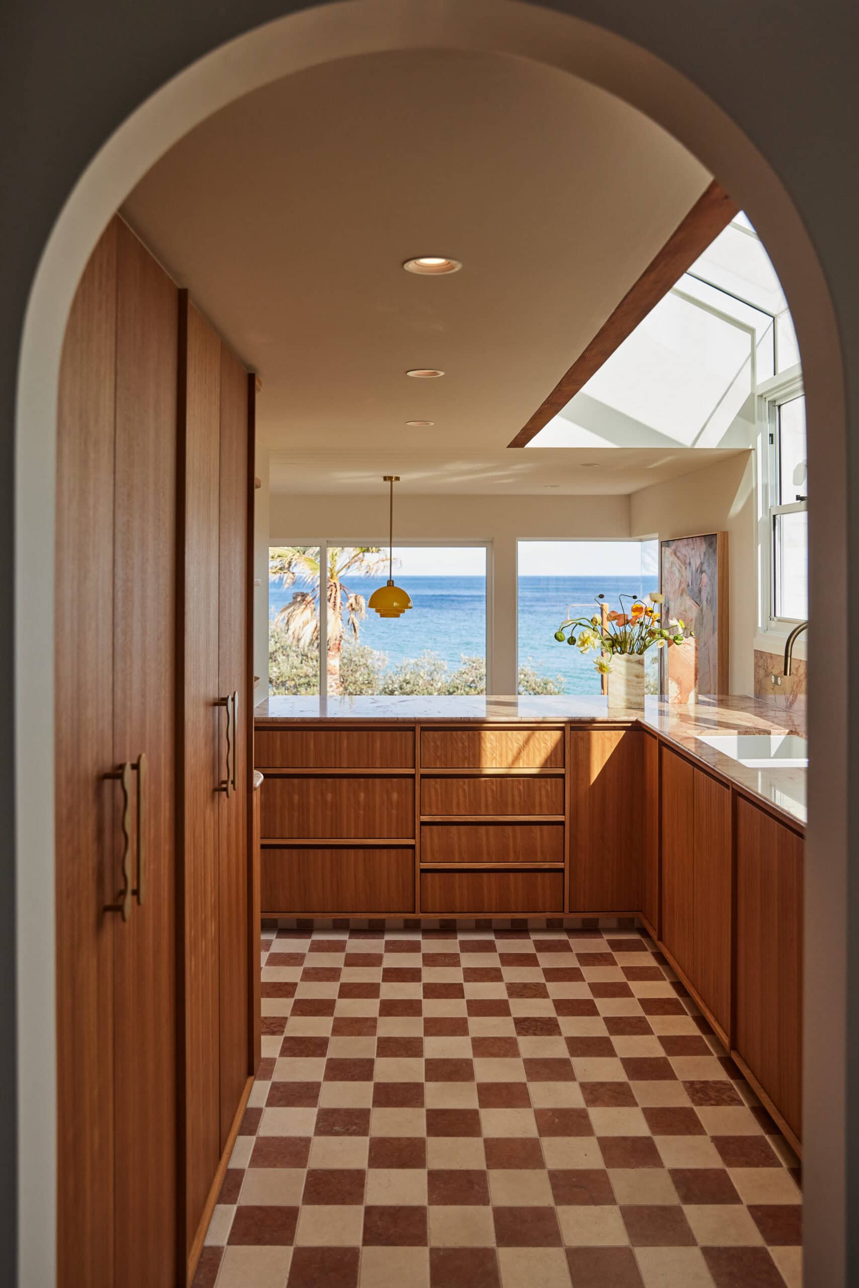 View through an arched wooden doorway into a galley kitchen with timber cabinetry, checkerboard tile flooring, a brass pendant light, and ocean views through the windows beyond.