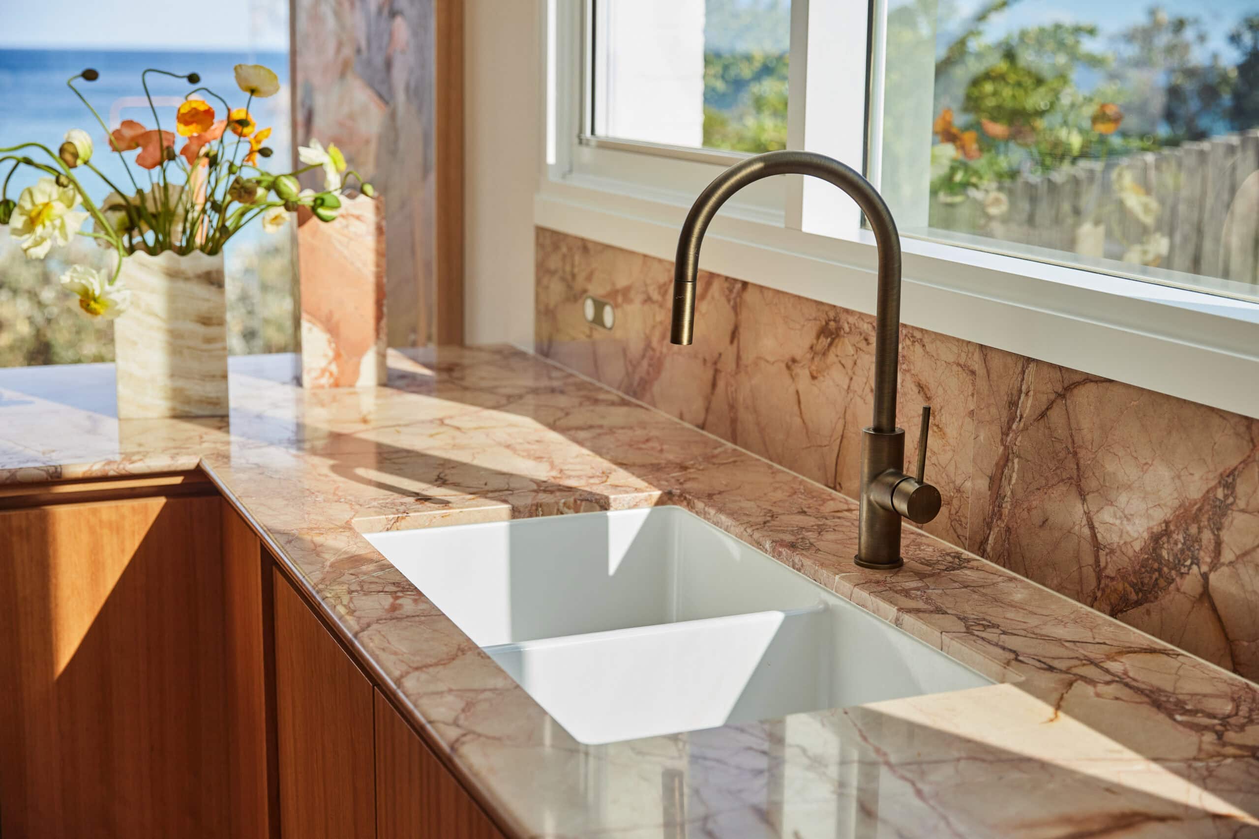 Close-up of a kitchen sink area with veined pink marble countertop and backsplash, a bronze gooseneck faucet, a white farmhouse sink, and a vase of poppies with an ocean view through the window.