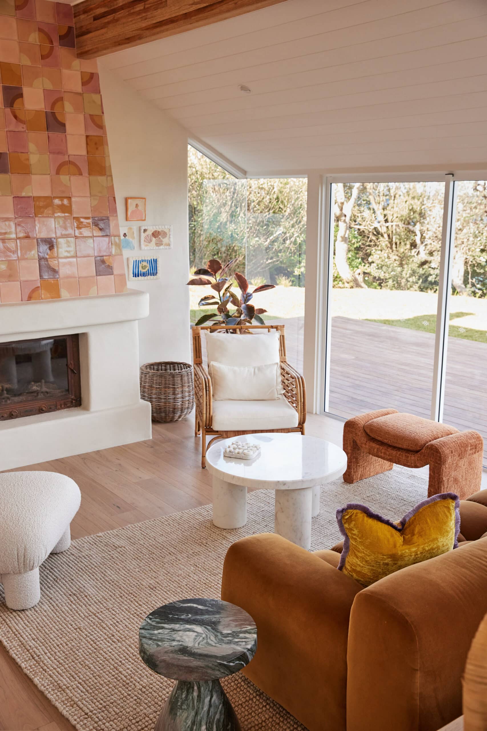 Coastal living room with a rust-colored velvet sofa, rattan armchair, marble side tables, and a handmade tiled fireplace, with sliding glass doors opening to a timber deck and garden.