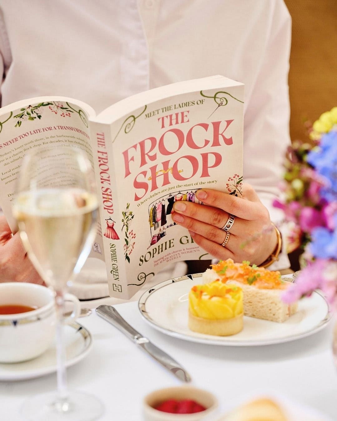 Person reading The Frock Shop by Sophie Green at a high tea table with champagne and finger sandwiches