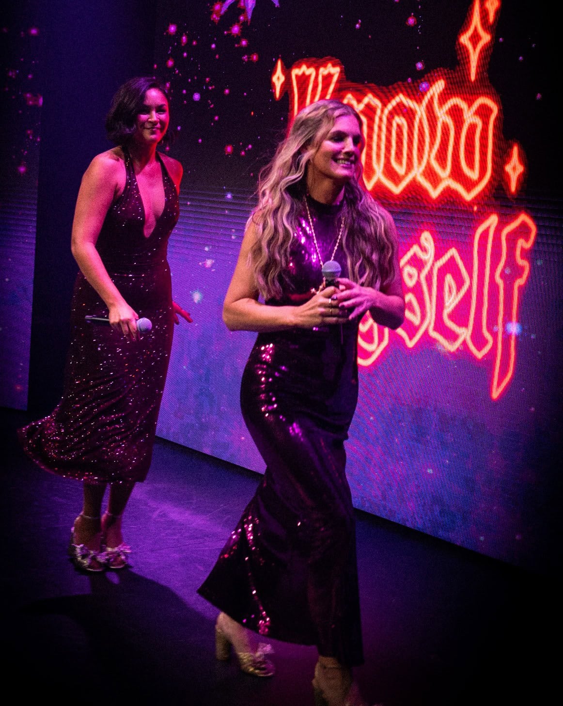 Two women on a stage performing at a live podcast event, both wearing sparkly sequined dresses in shades of purple/magenta and holding microphones. Behind them, a large screen displays glowing neon-style lettering reading 