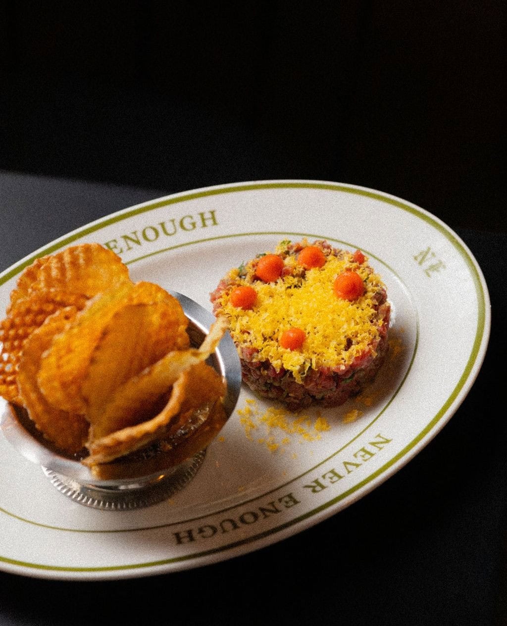 Beef tartare with egg yolk shavings on a white plate served with a silver dish of crimped crisps.