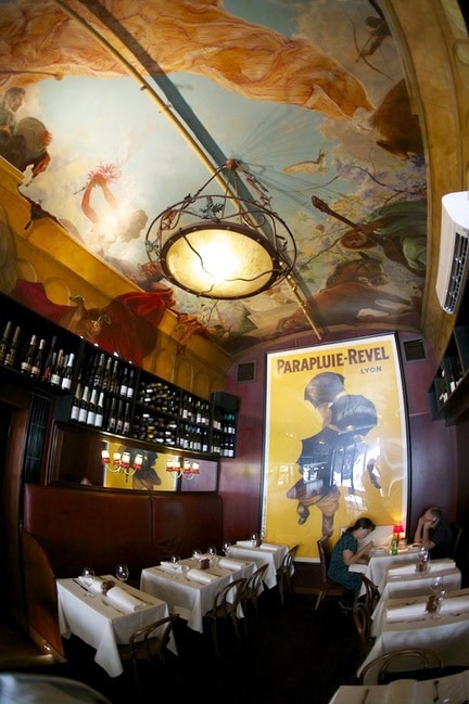 Interior of Bistrot d’Orsay on Collins Street in Melbourne, with white tablecloths, dark timber, a painted ceiling and vintage French artwork.