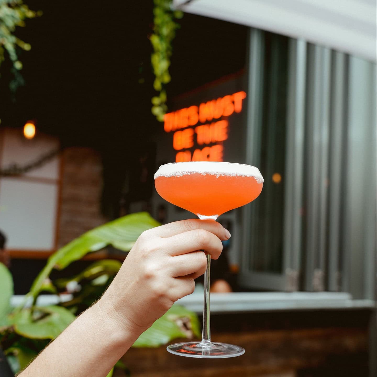 A hand holding a orange-oink cocktail in front of an LED sign saying 