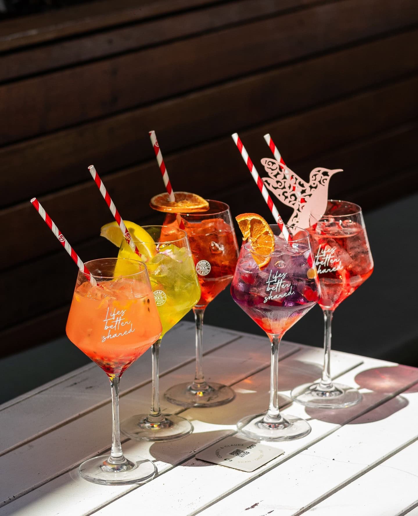Five brightly coloured cocktails in spritz glasses, garnished with lemon wedges or dried orange on a white sunny table.
