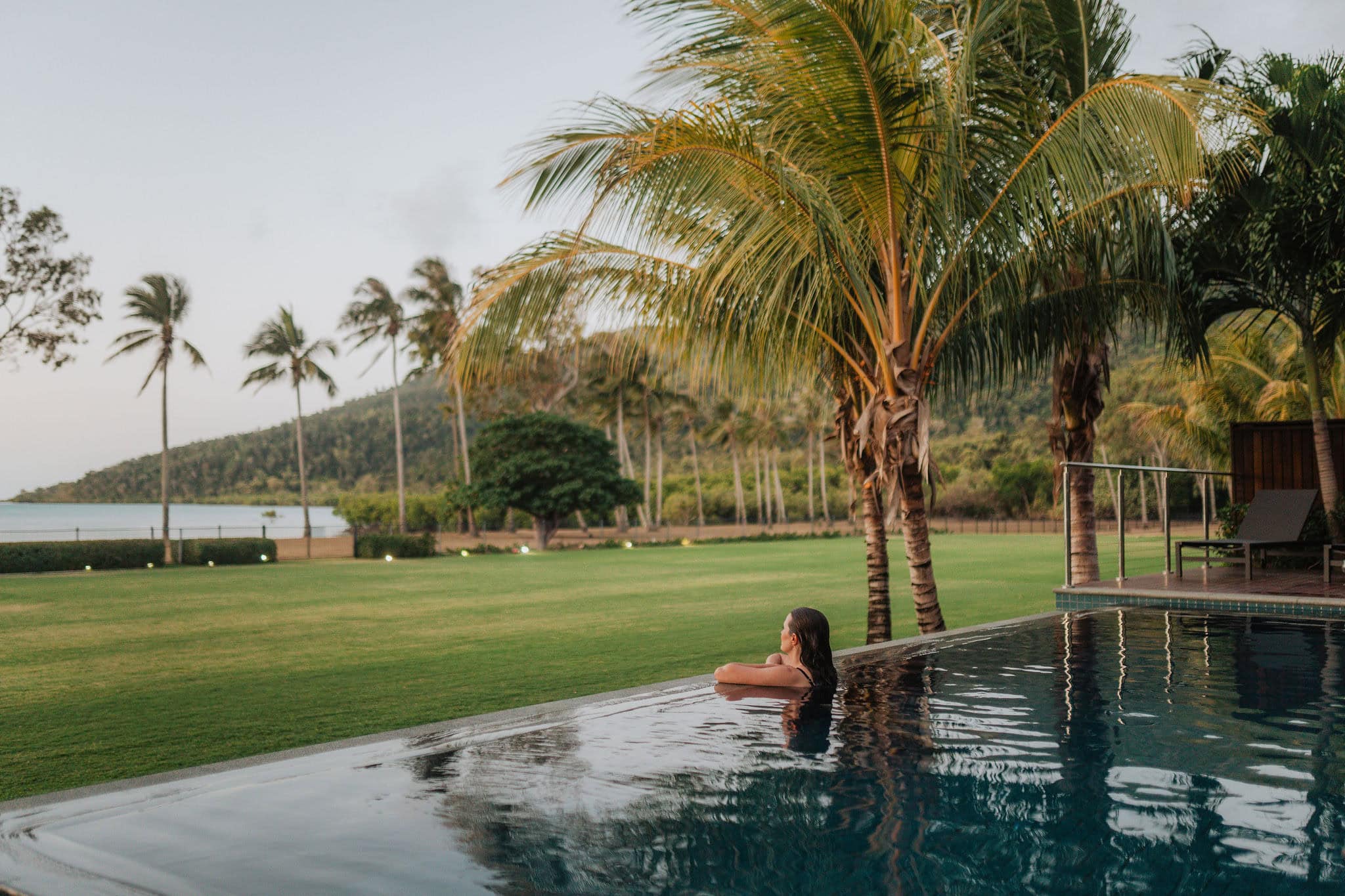 Guest relaxing in an infinity pool framed by palm trees, lawns and the waterfront at Yangaro, Funnel Bay.