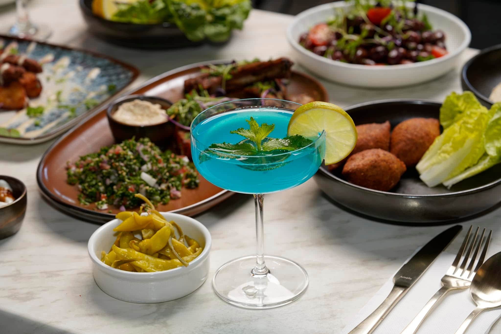 A bright blue cocktail in front of five sharing plates of Middle Eastern food.