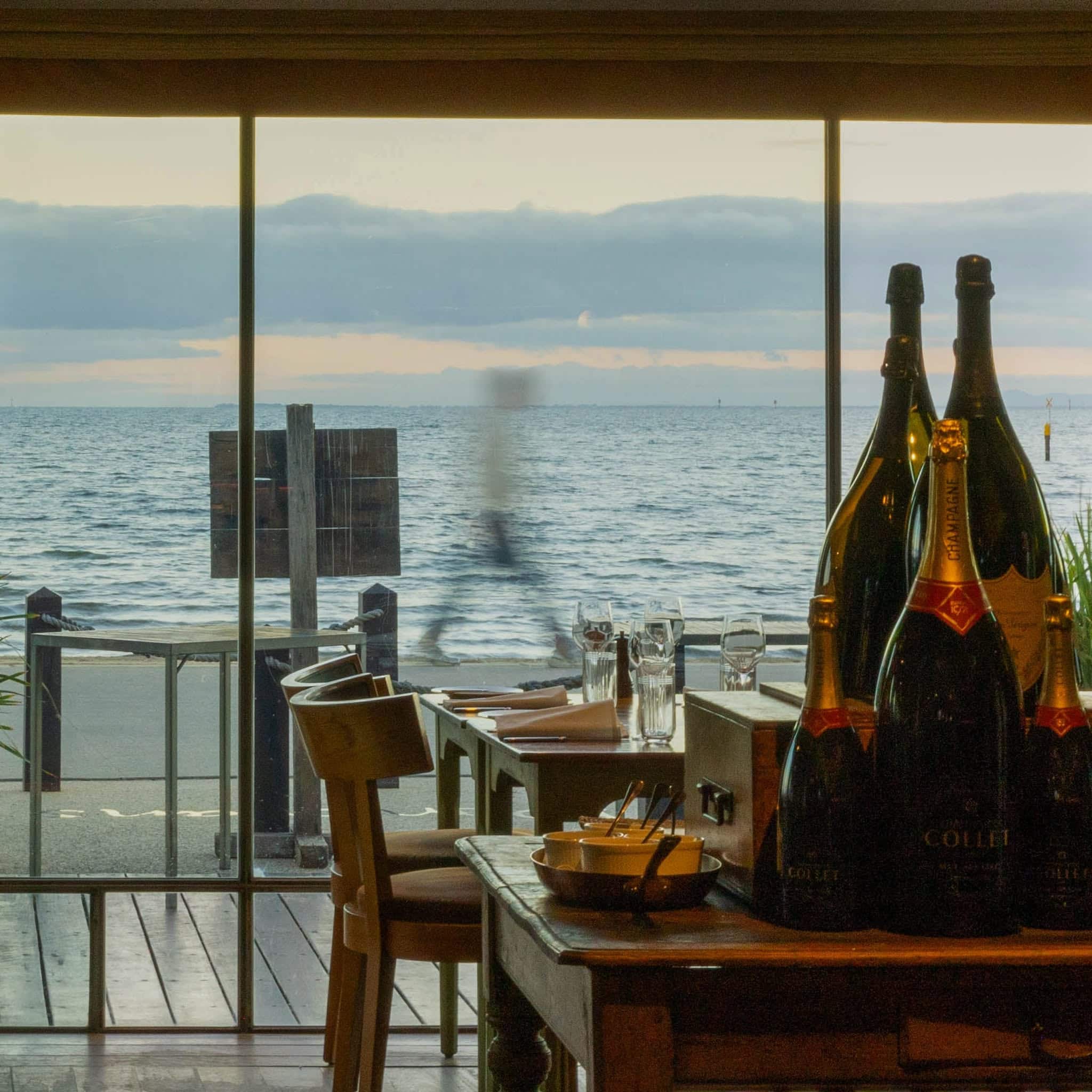 Dining room at Donovans in St Kilda overlooking Port Phillip Bay, with Champagne bottles and waterfront tables at sunset.