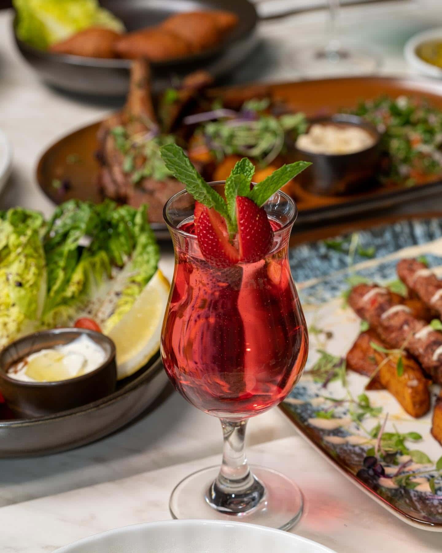 A red sparkling fruit drink in the foreground of a table, with share plates on the table behind it.