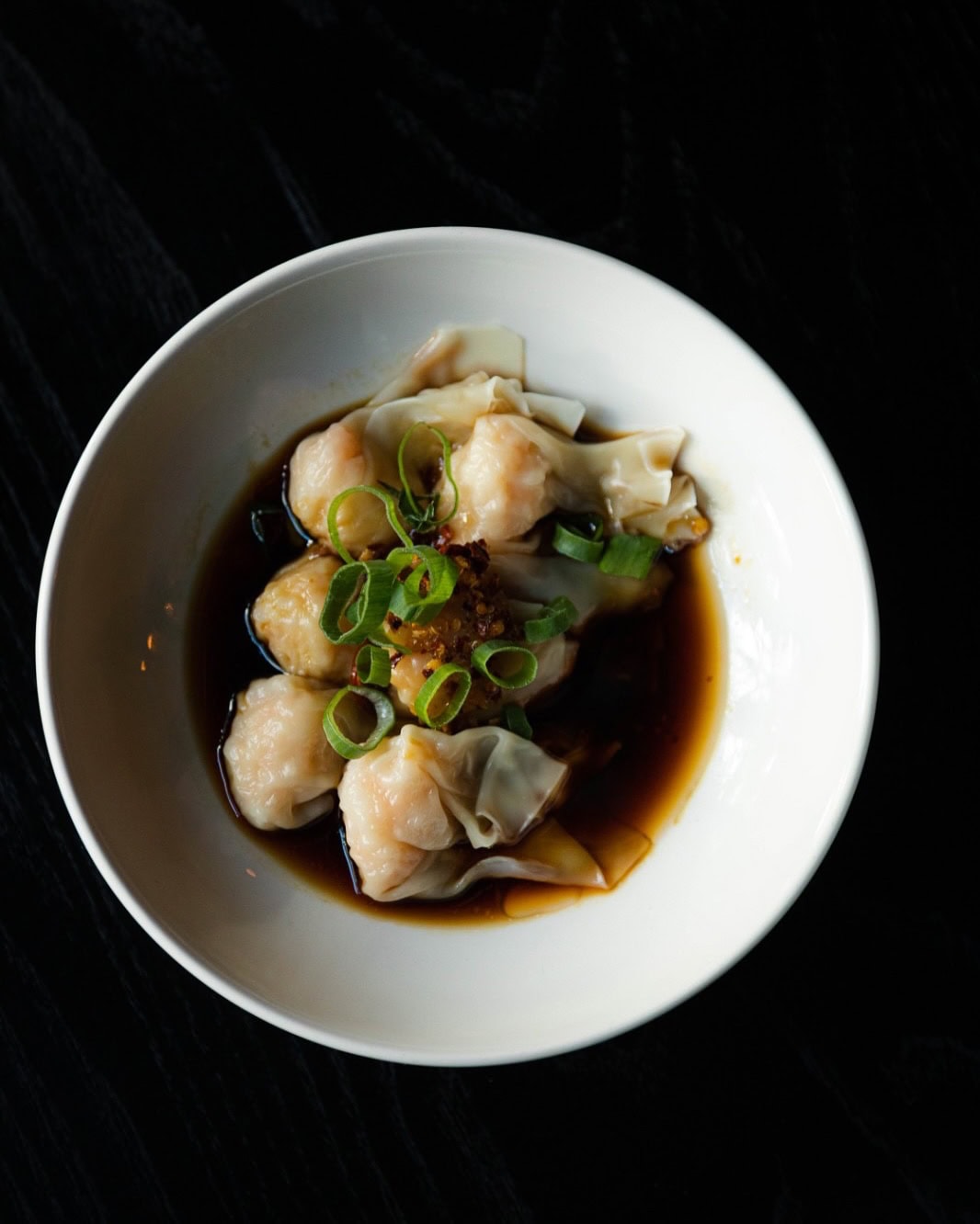 A soupy bowl filled with dumplings and garnished with spring onions on a black table.