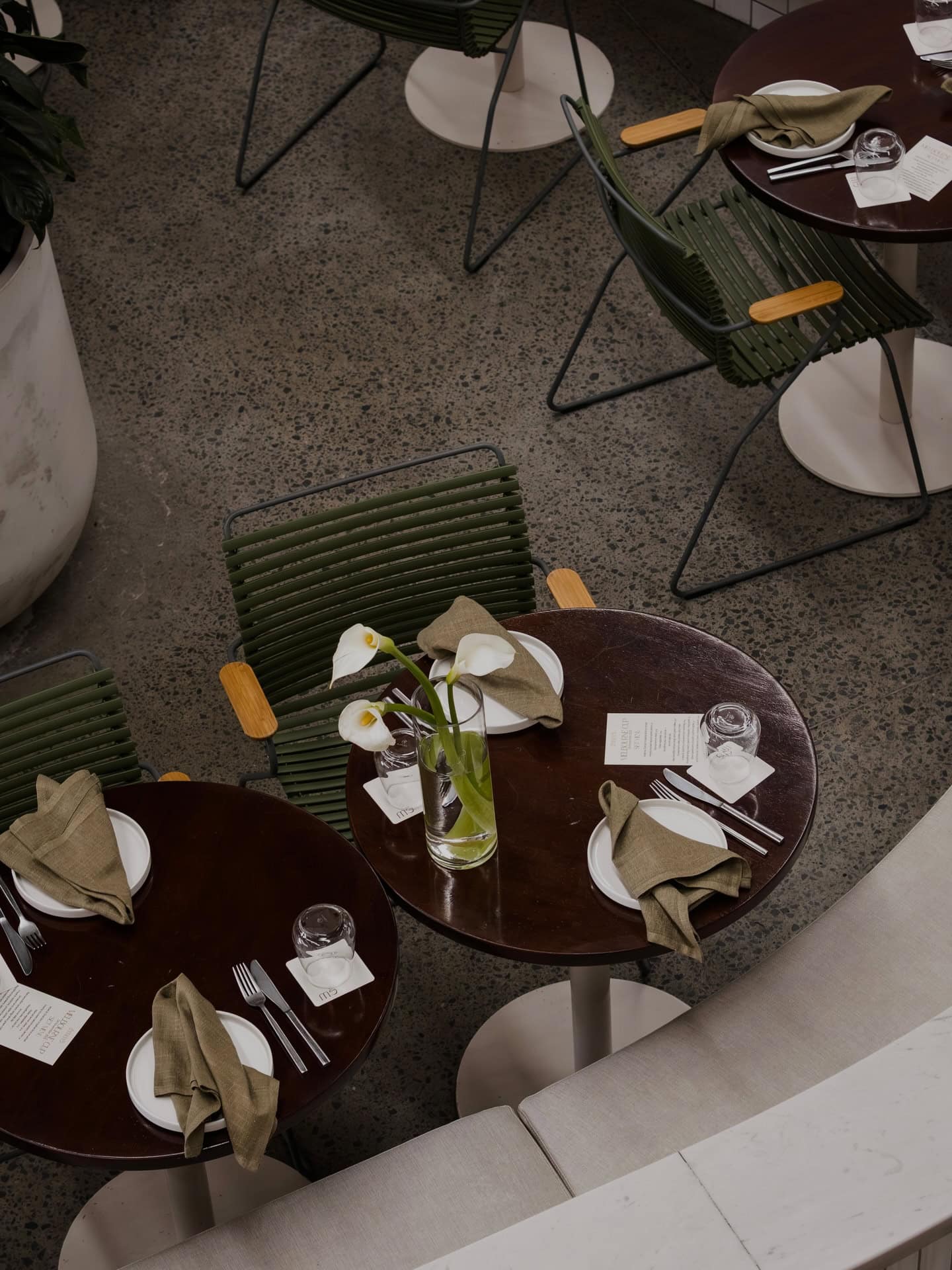A top-down shot of dark brown outdoor dining tables, set with white plates, cutley and sage cloth napkins.