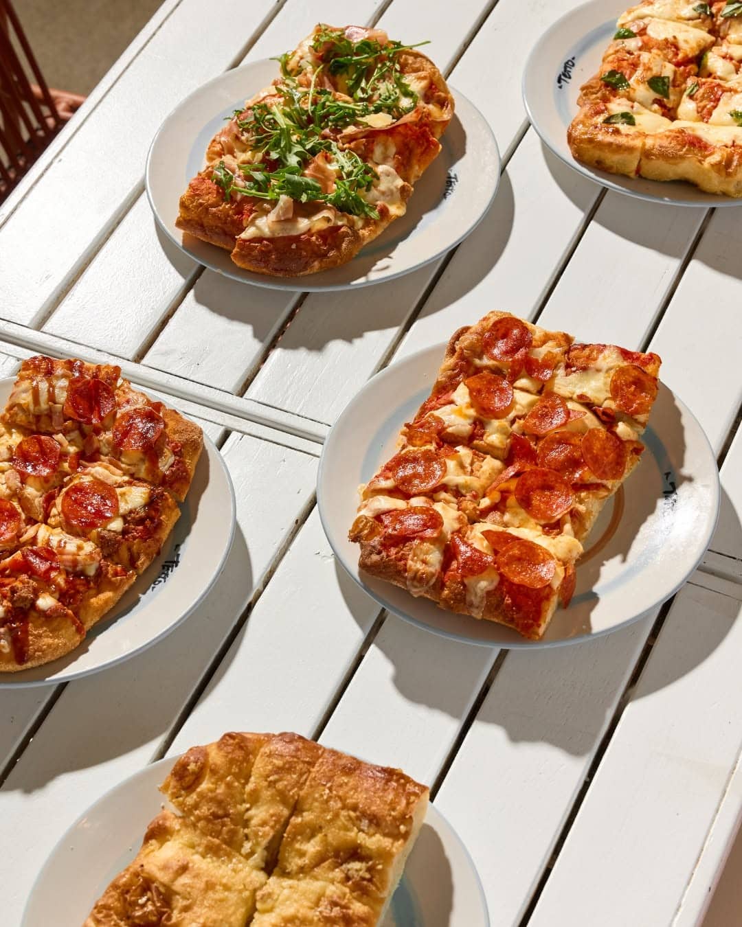 Five plates of focaccia on a white table in the sunshine, topped with pepperoni, cheese, rocket, garlic.