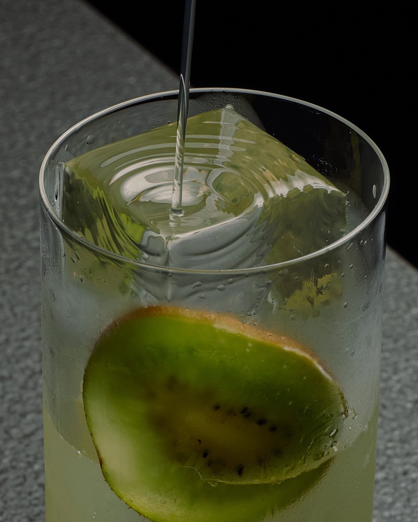 A macro shot of a highball being poured, with a large long ice cube in the glass and a slice of kiwi wedged against the inside.