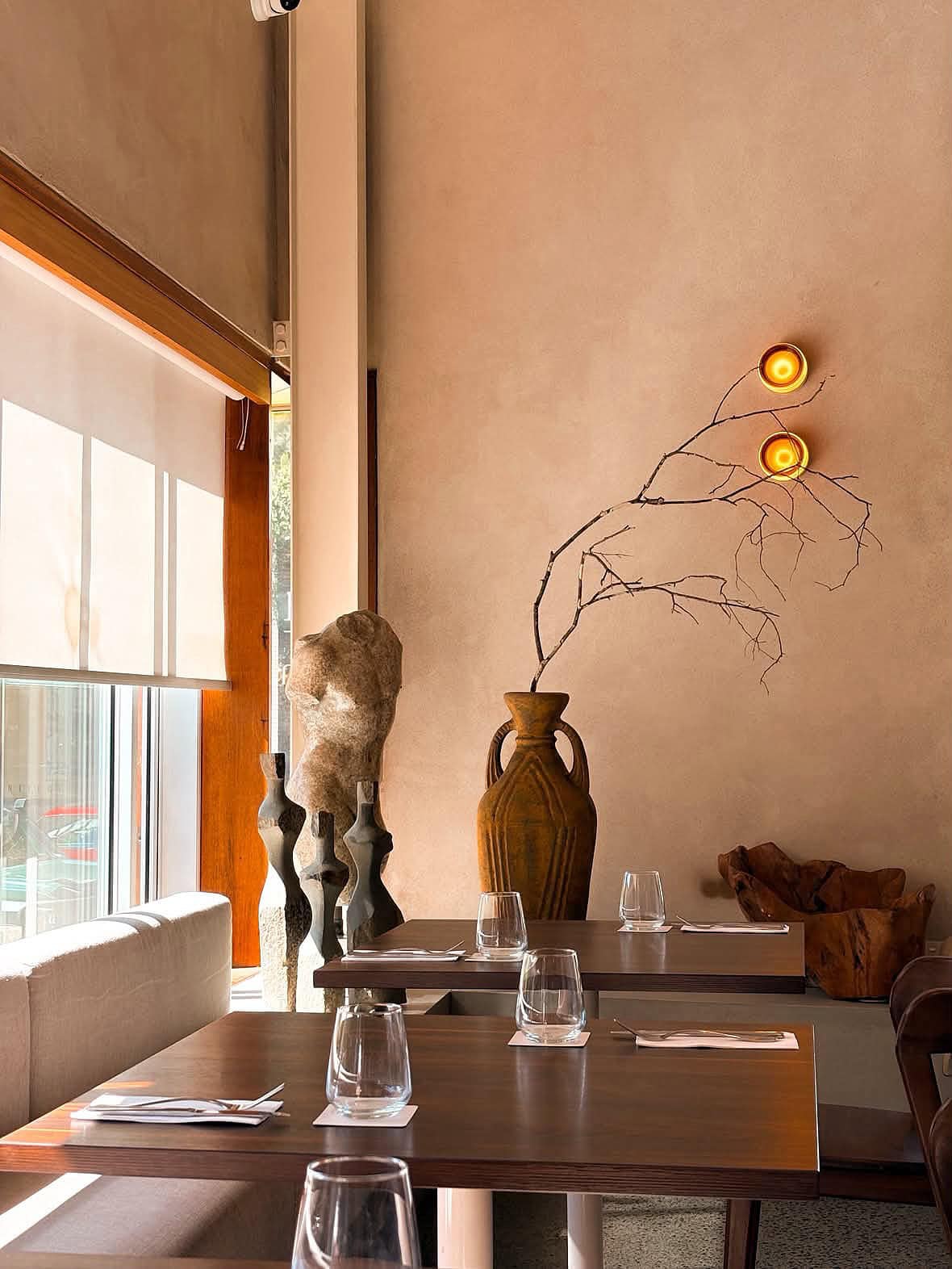 A warm-toned restaurant interior with wooden tables, a vase with a dried branch and sculptures.