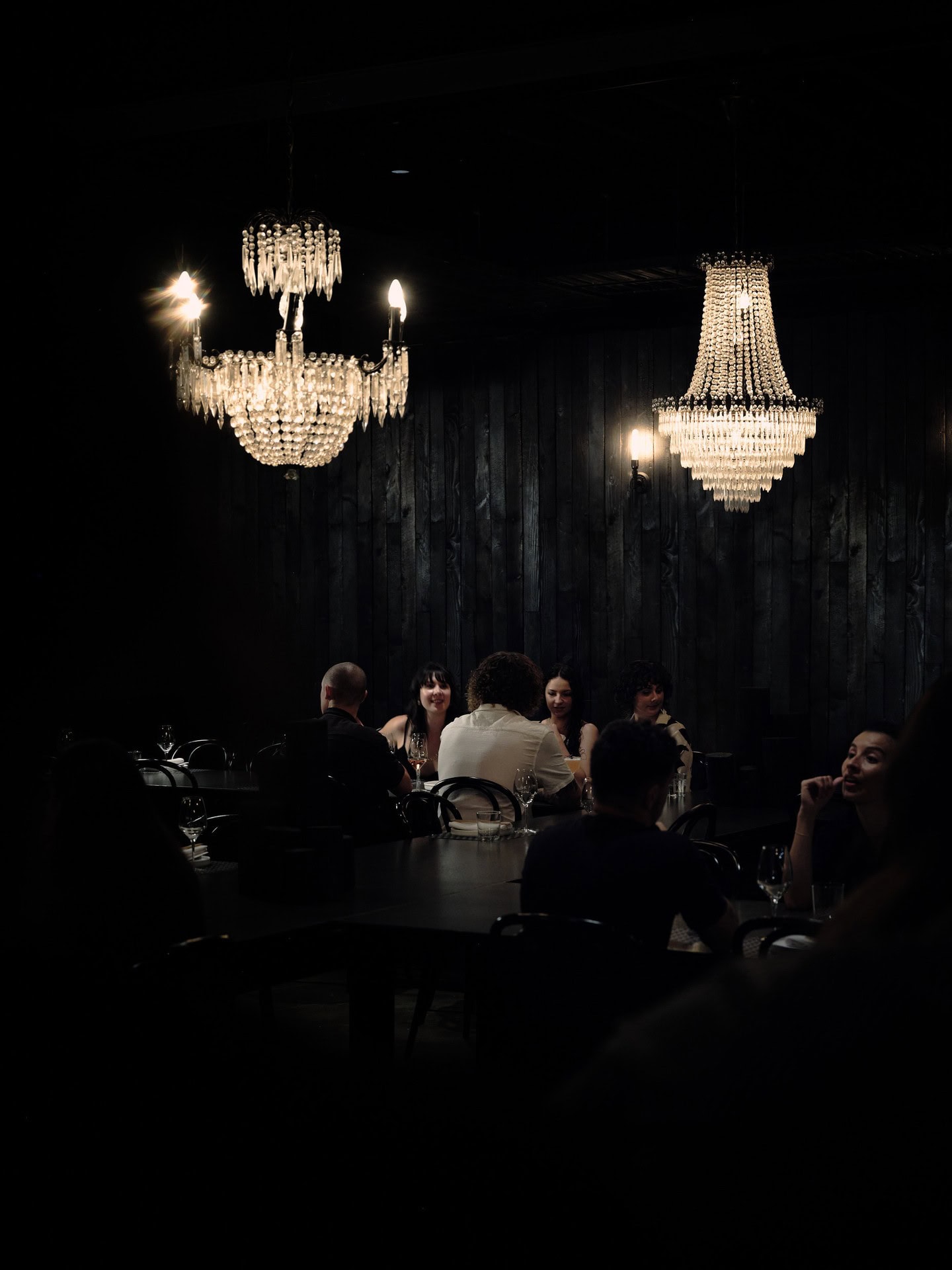 A dimly lit room with two different crystal chandeliers, illuminating diners at black tables below.