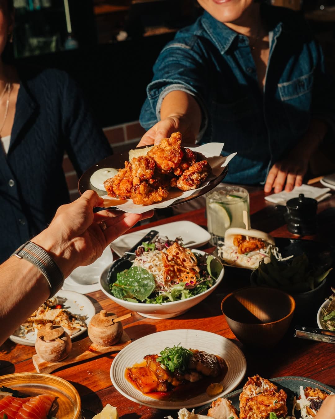 A smiling person passes a full plate of karaage chicken to a fellow diner, over a table laden with share plates and cocktails.