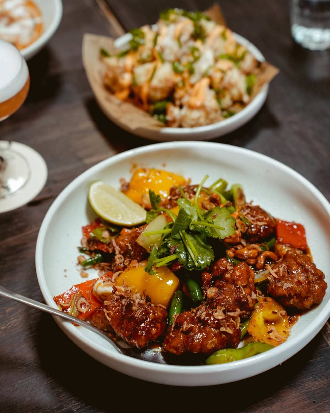 A white bowl of chicken cashew stirfry in the foreground, on a brown table.