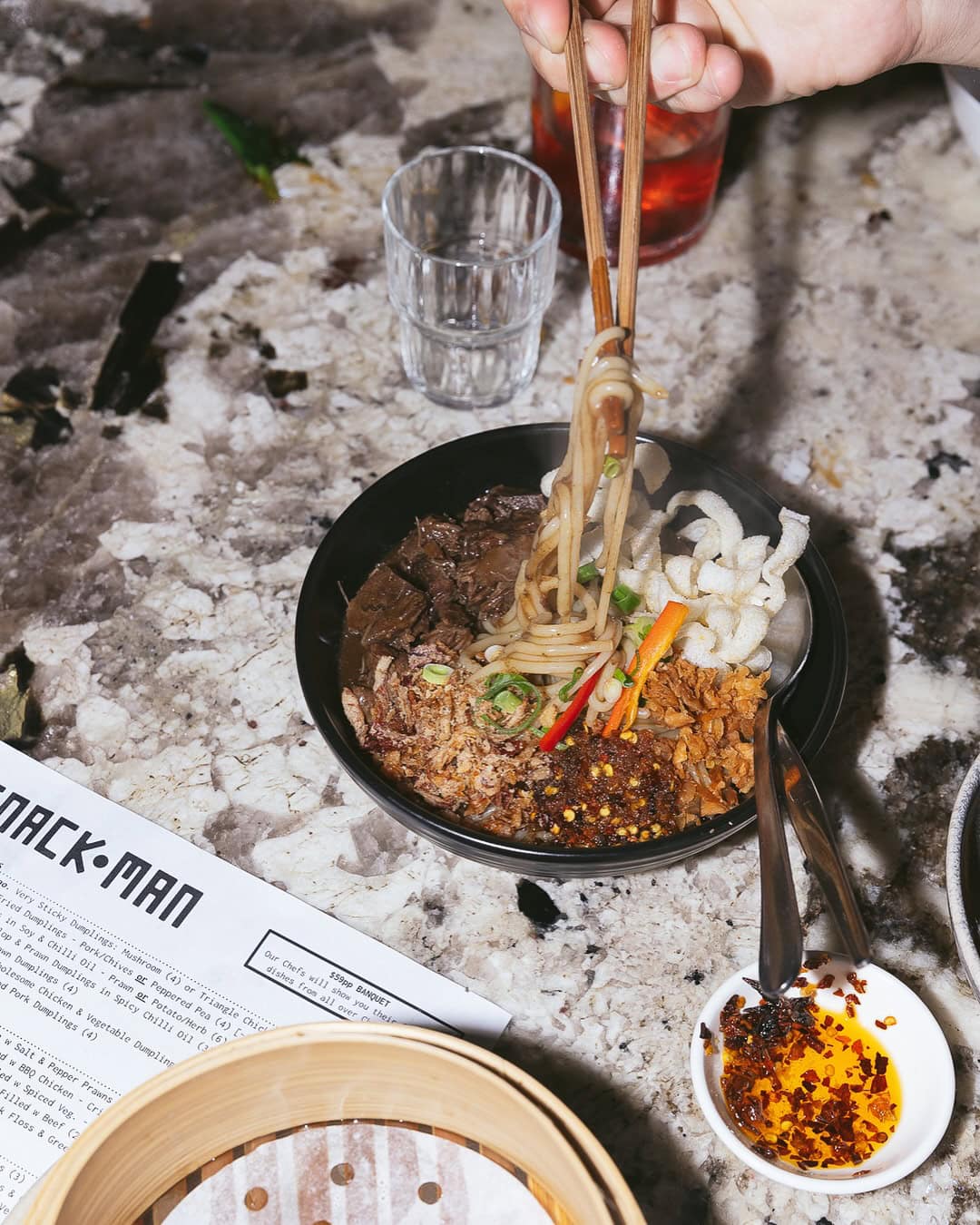 Chopsticks are pulling noodles from a bowl full of beef, vegetables and garnish.