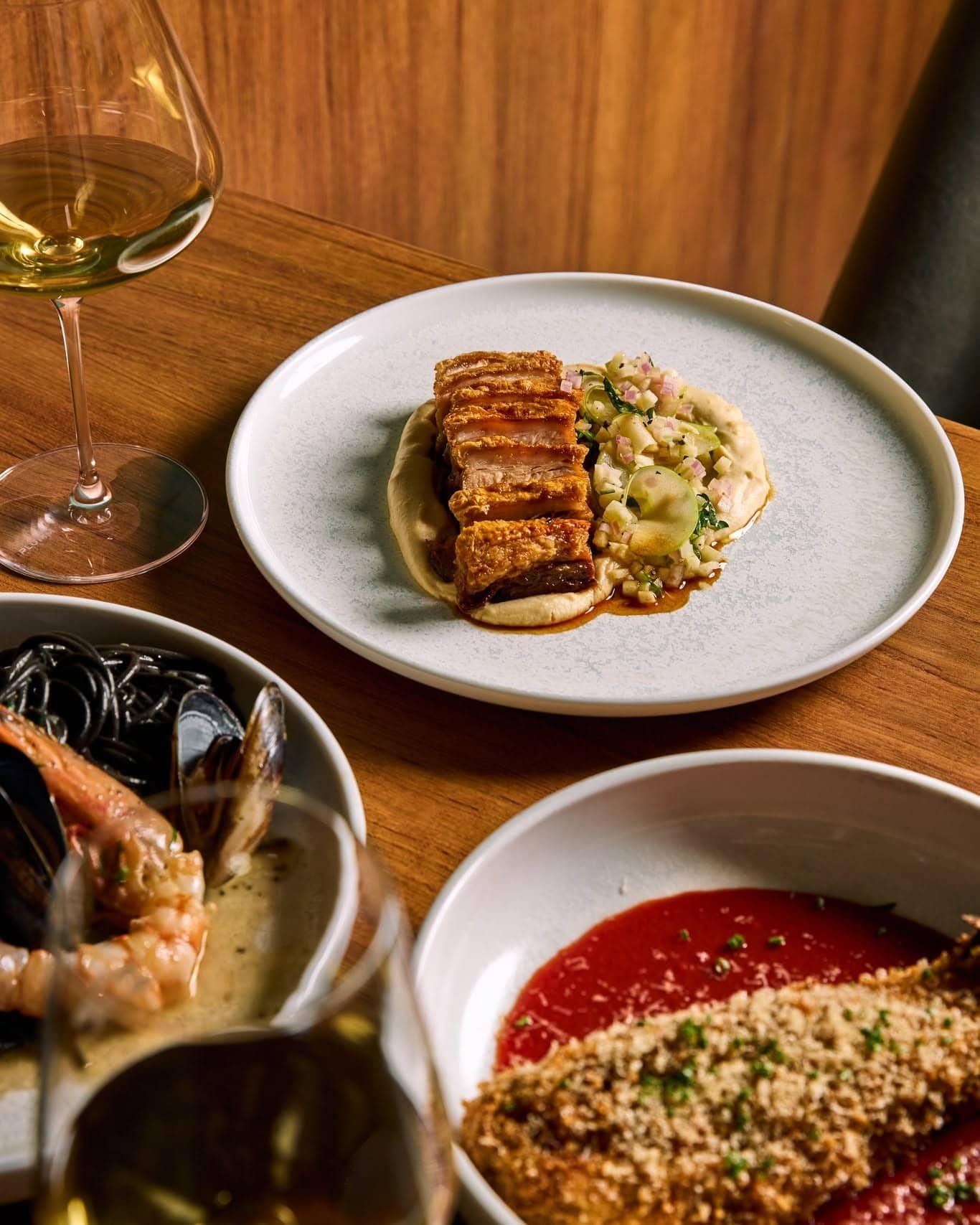 Pork Belly, Crumbed Eggplant & Squid Ink Pasta served on white plates at a cosy wine bar.