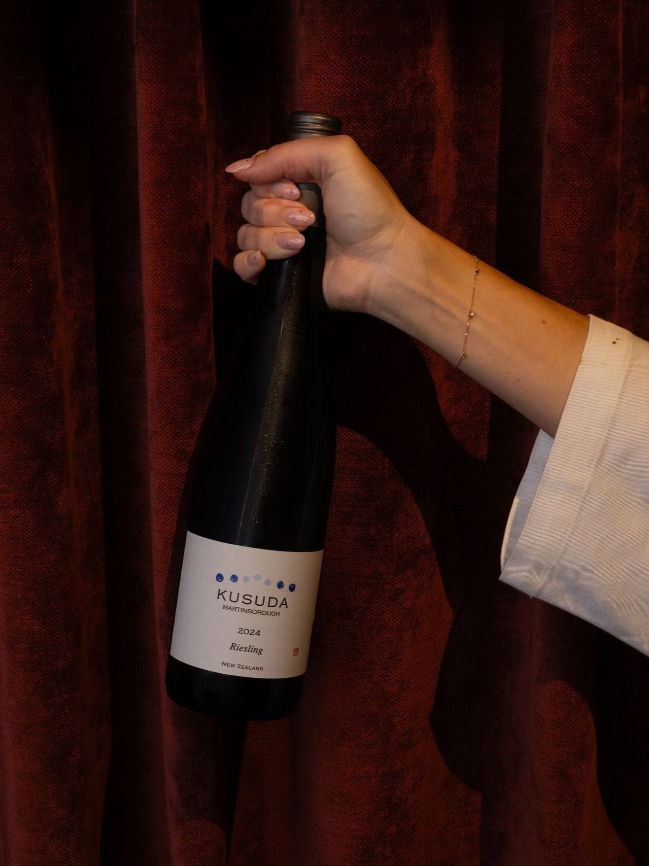 A hand holding a bottle of 2024 Kusuda Riesling in front of a heavy red curtain