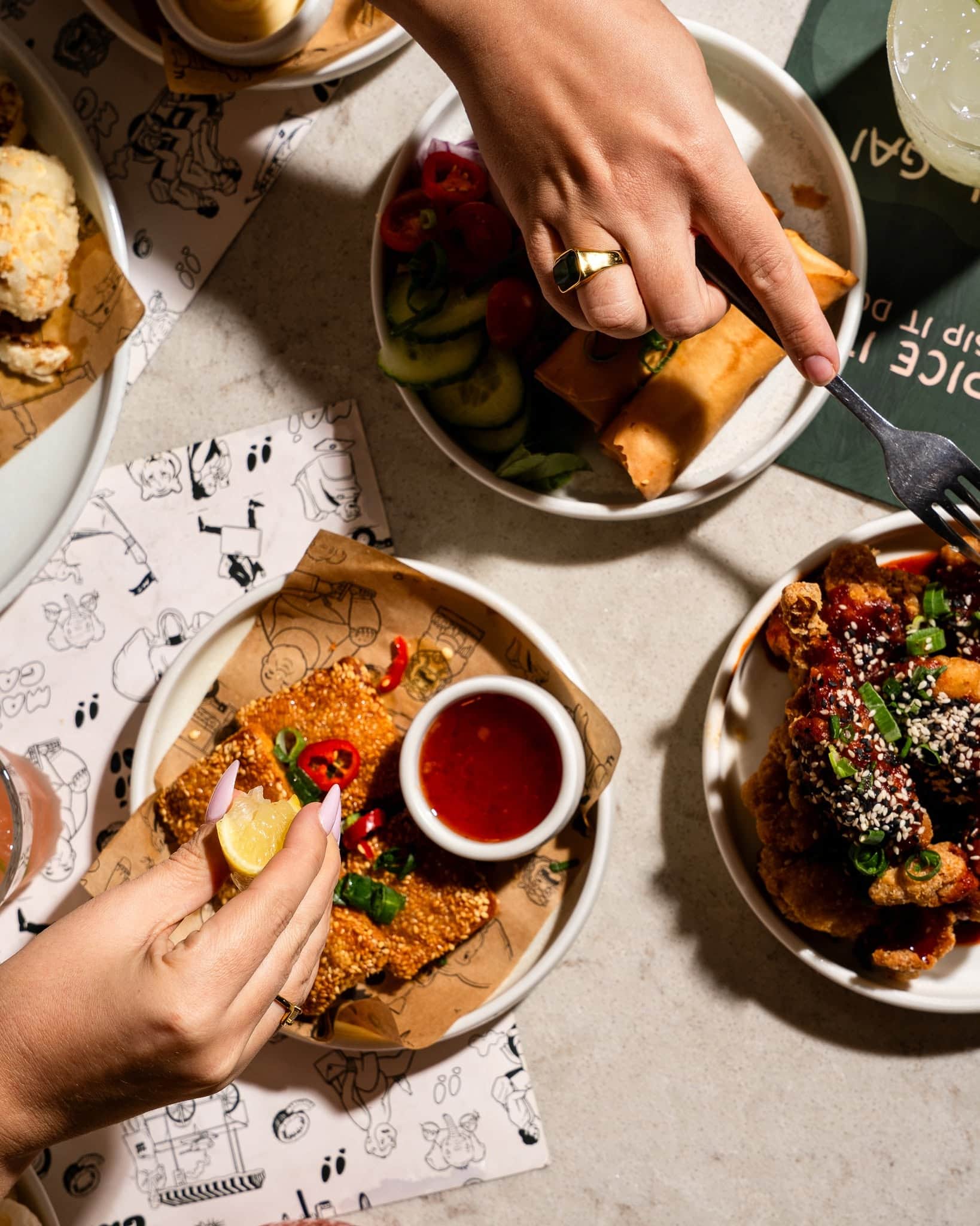 Two hands reaching across the table, one about to spear some food on a fork, one squeezing a lemon wedge.