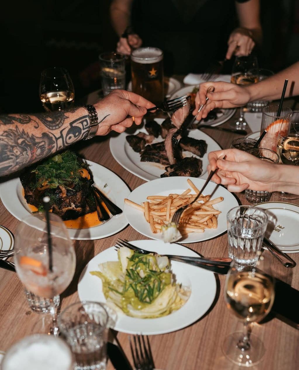 Three people's hands reach across a table loaded with share plates of steak, fries, salad, wine glasses and cocktails.