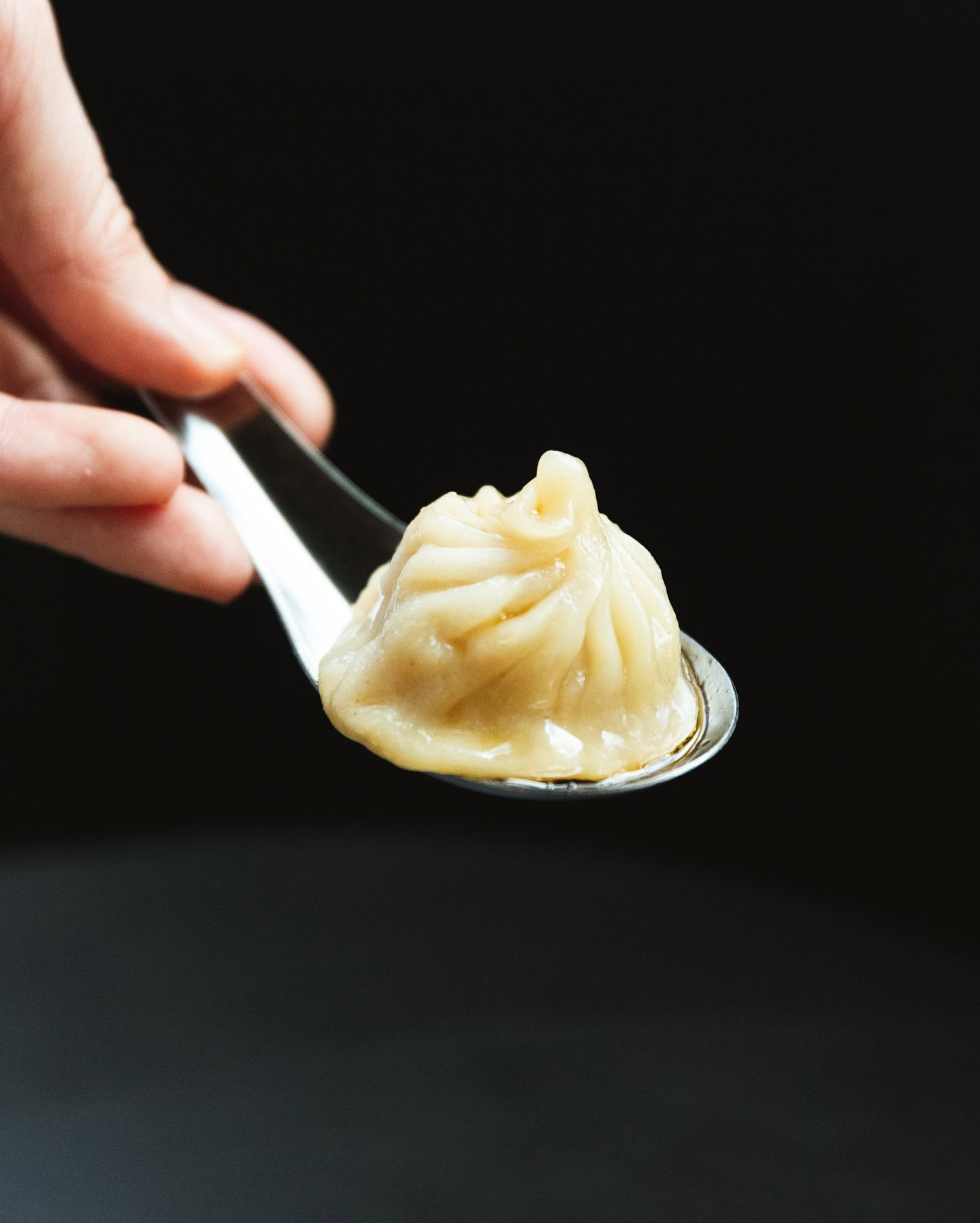 A xiao long bao soup dumpling on a spoon, held suspended in the air with a black background.